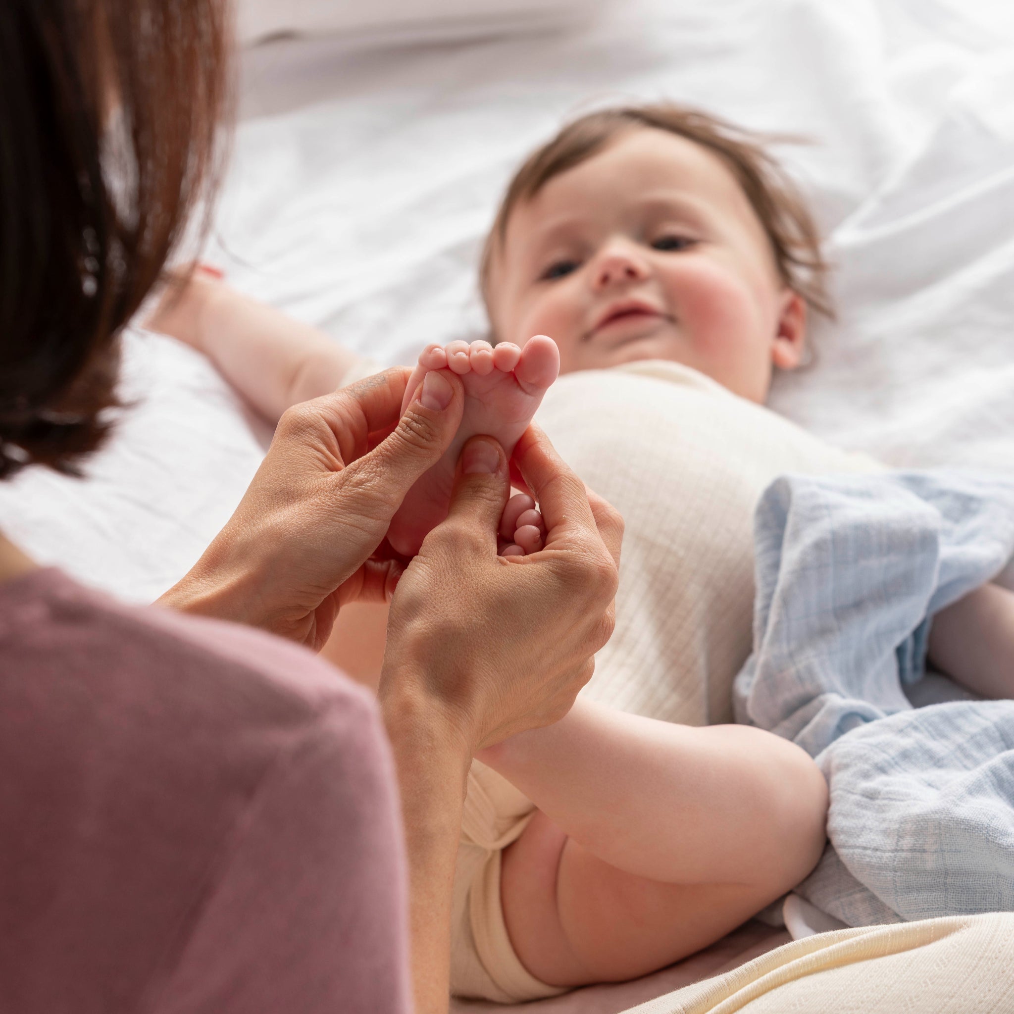 Eine Frau massiert sanft den Fuß eines lächelnden Babys, das in einem mit weißen Laken bedeckten Bett liegt. Das Baby schaut zu der Frau auf und ist in eine hellblaue Decke eingewickelt.