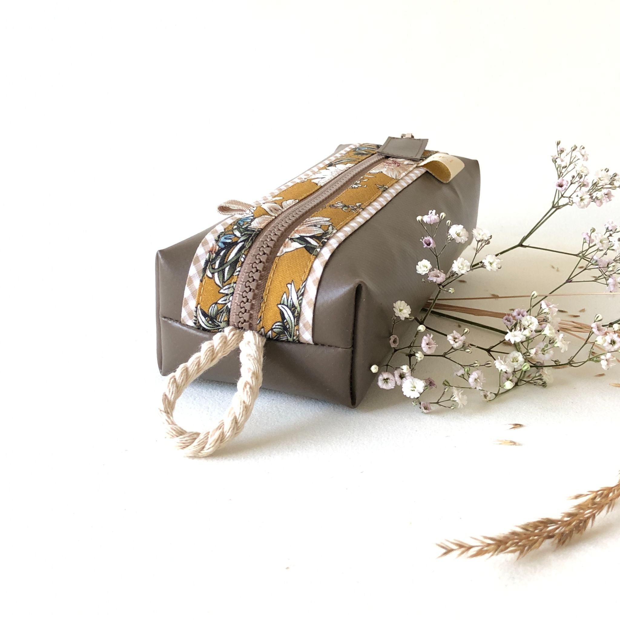 Eine rechteckige braune Tasche mit einem Seilgriff und einem Reißverschluss, mit einem dekorativen gelben und floralen Stoffstreifen, neben zarten Zweigen kleiner weißer Blumen auf hellem Hintergrund.