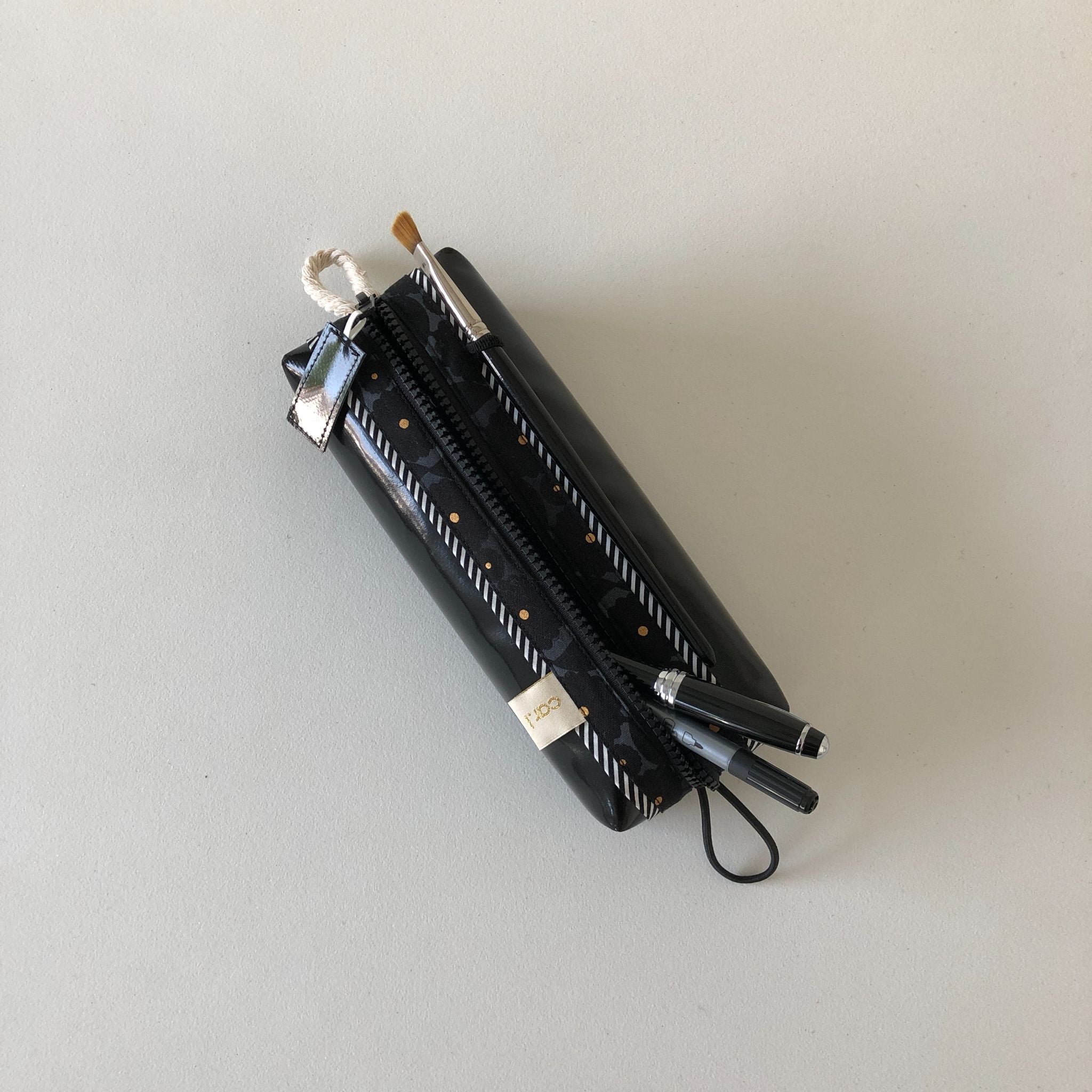 Ein schwarzes Mäppchen mit weißen diagonalen Streifen und Punkten liegt auf einer hellen Fläche, teilweise geöffnet, und aus dem Reißverschluss ragen ein Pinsel und zwei Stifte.