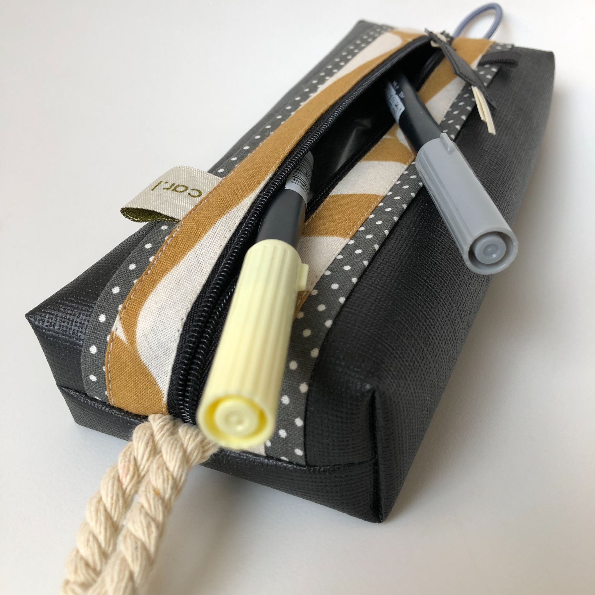 Ein schwarz-gelbes, rechteckiges Mäppchen mit Reißverschluss, grauen Tupfen und gelben Streifen und einem Seilgriff, in dem zwei Stifte - ein grauer und ein gelber - zum Teil herausragen.