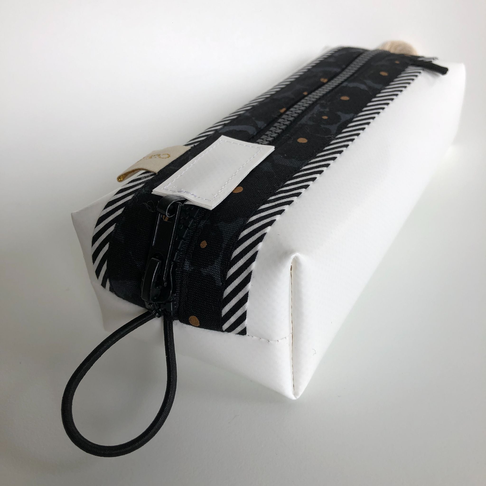 Eine weiße rechteckige Tasche mit Reißverschluss und einem schwarz-weiß gemusterten Stoffstreifen um den Reißverschluss herum, mit einer kleinen elastischen Schlaufe an einem Ende und goldenen Punkten auf dem Stoff.