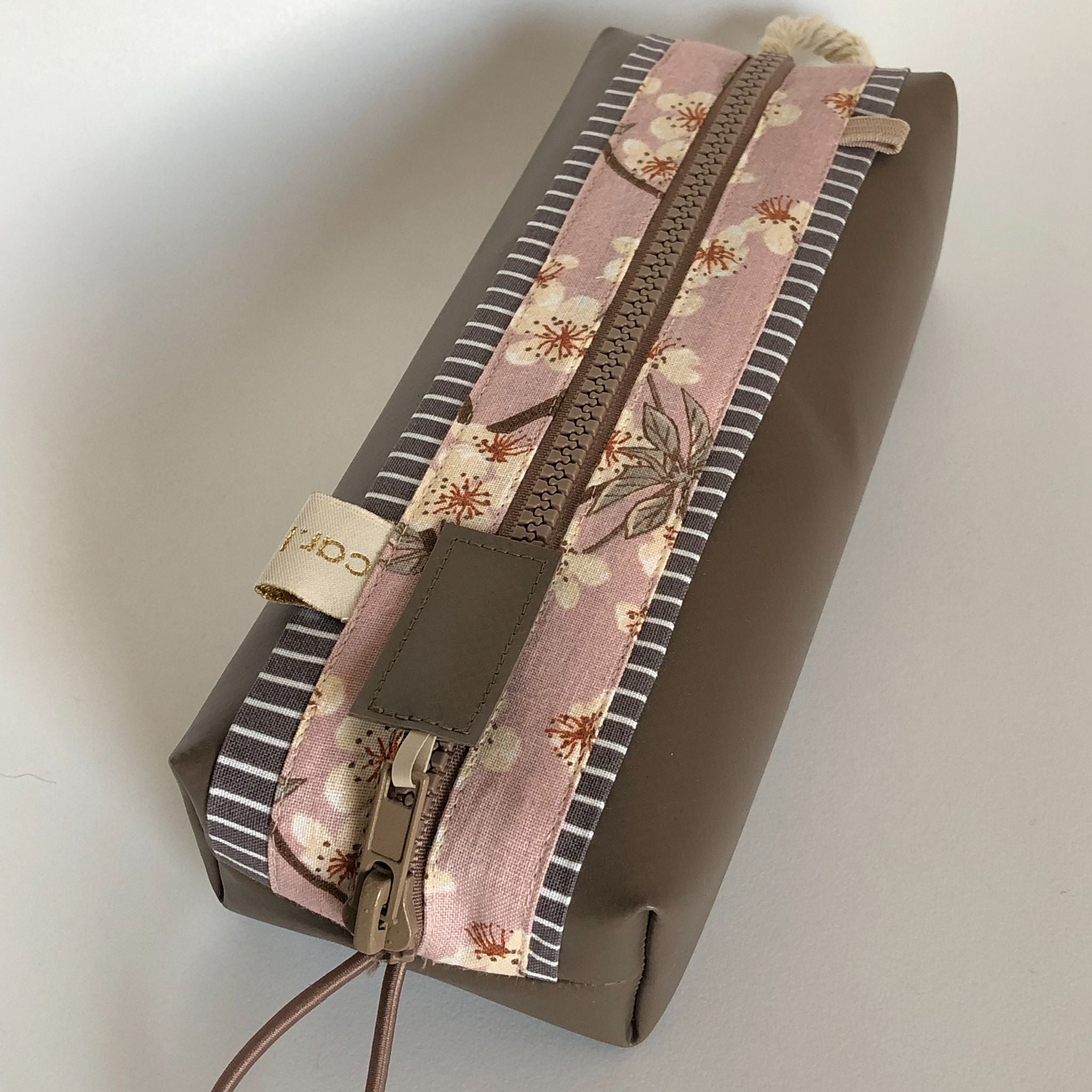 Eine rechteckige, braune Tasche mit Reißverschluss mit einem rosafarbenen, geblümten Stoffstreifen und grauen, gestreiften Rändern auf der Oberseite, die auf einer hellen Oberfläche ruht. Der Reißverschluss hat eine kleine Zuglasche aus Leder.