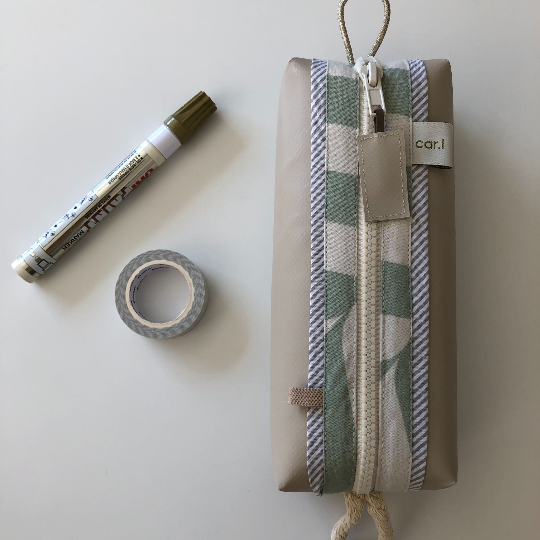 Ein goldener Marker, eine Rolle graues Washi-Tape und ein rechteckiges beiges Federmäppchen mit Reißverschluss und Zierstreifen sind auf einer weißen Fläche angeordnet.