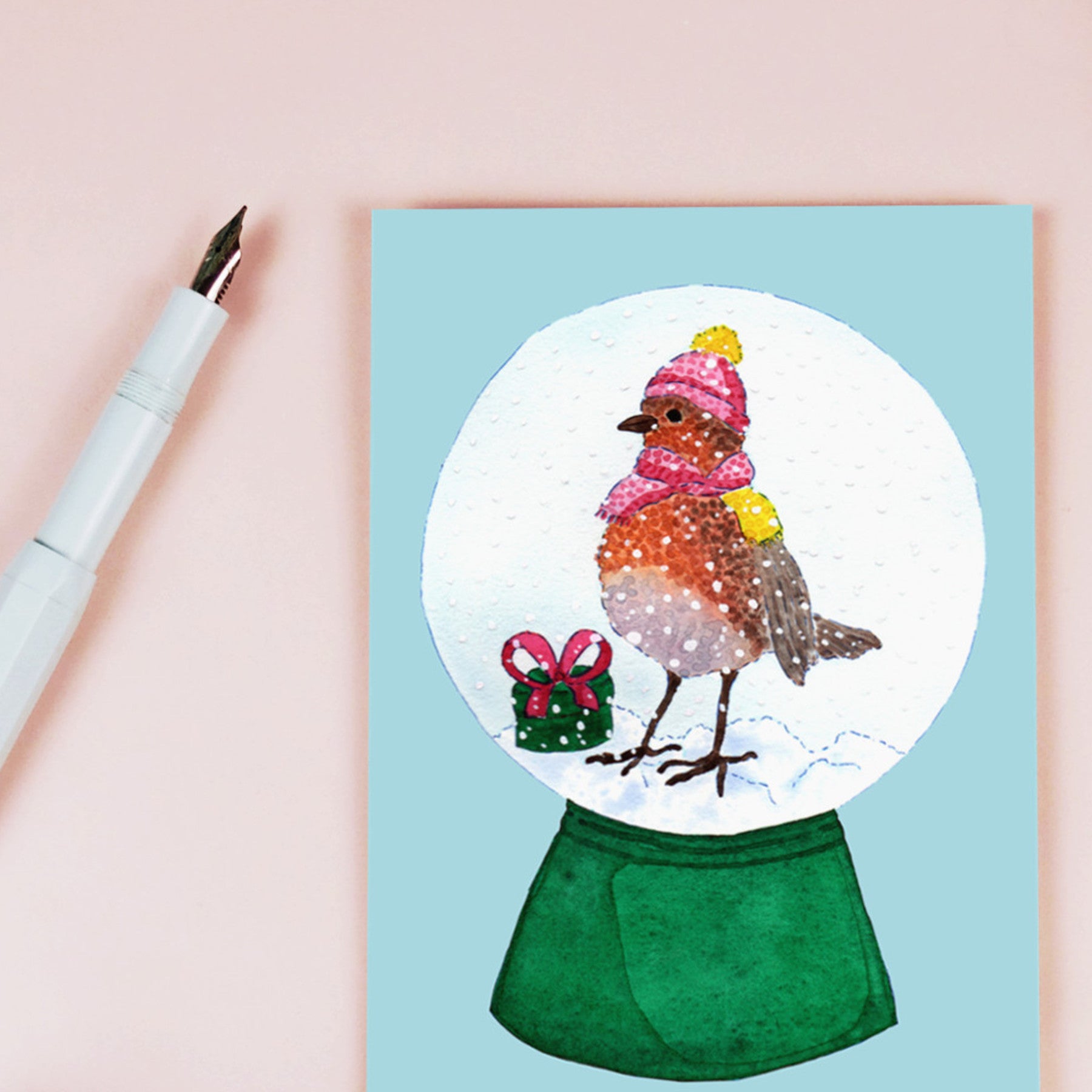 Eine Grußkarte mit der Illustration eines Rotkehlchens mit rosa Hut und Schal in einer Schneekugel, das neben einem kleinen grünen Geschenk mit einer roten Schleife steht. Ein Stift liegt neben der Karte auf einer blassrosa Fläche.