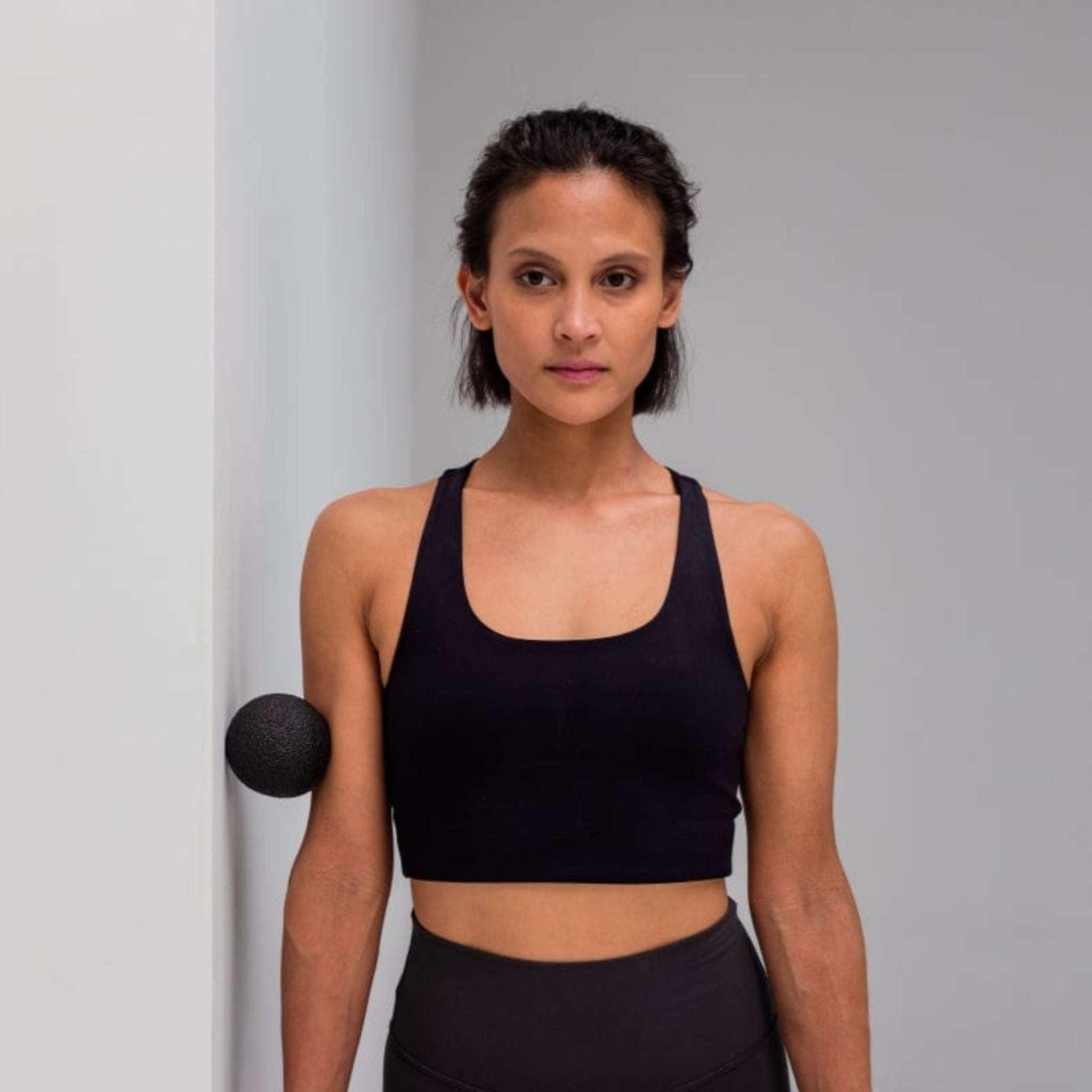 Eine Frau in Sportkleidung lehnt an der Wand, wobei ein Massageball zwischen Wand und Arm eingeklemmt ist.