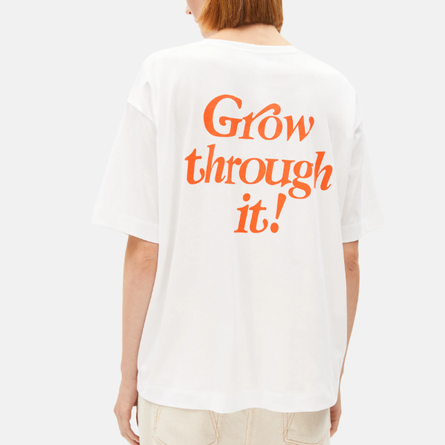 Eine Person mit kurzen, hellbraunen Haaren trägt ein weißes T-Shirt mit orangefarbener Aufschrift auf dem Rücken, die lautet: "Wachsen Sie durch!" Die Person wird von hinten gezeigt und trägt ebenfalls eine helle Hose.