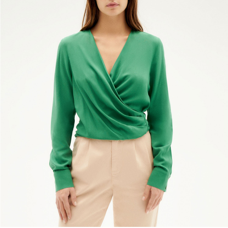 Eine Frau mit langen braunen Haaren trägt eine grüne Wickelbluse mit langen Ärmeln und eine beigefarbene Hose. Sie steht vor einem schlichten hellen Hintergrund und schaut mit neutralem Blick direkt in die Kamera.