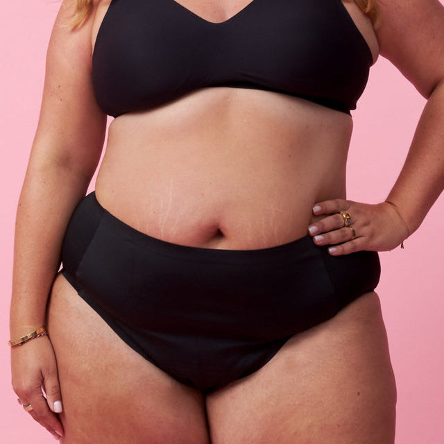 Eine Frau mit gewelltem Haar, die einen schwarzen Bikini trägt, steht vor einem rosafarbenen Hintergrund und zeigt sichtbare Dehnungsstreifen und Schmuck an ihrer Hand und ihrem Handgelenk.