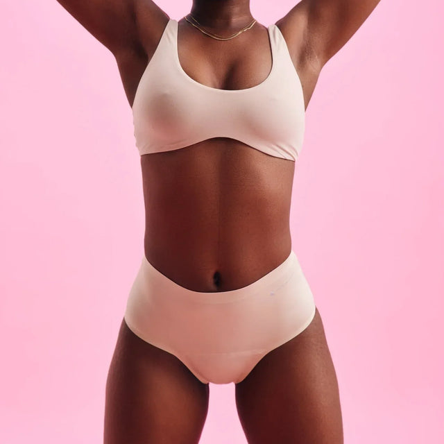 Eine Frau steht selbstbewusst vor einem rosafarbenen Hintergrund und trägt einen passenden nudefarbenen BH und hochtaillierte Unterwäsche. Ihre Arme sind erhoben und ruhen hinter ihrem Kopf. Sie blickt direkt in die Kamera.