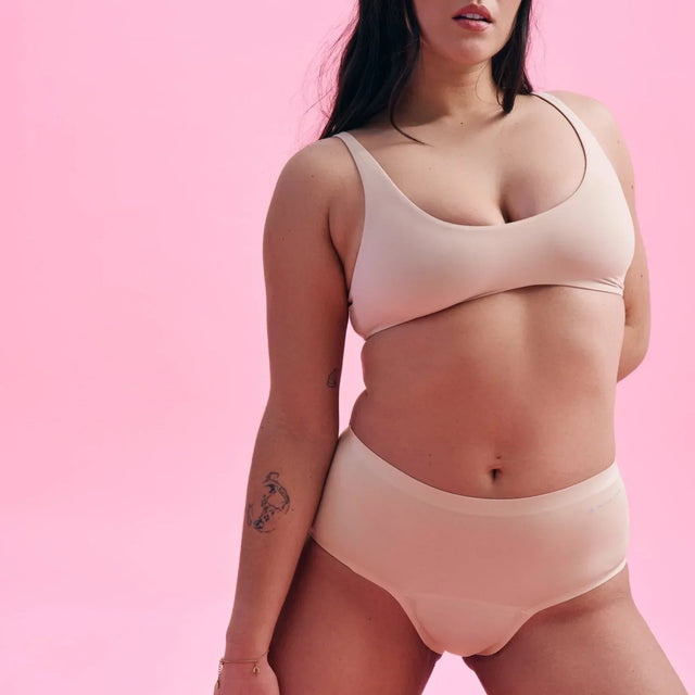 Eine Frau mit langen dunklen Haaren posiert selbstbewusst in nudefarbener Unterwäsche vor einem kräftigen rosa Hintergrund. Sie hat einen Arm hinter dem Rücken und sichtbare Tattoos auf dem linken Arm.