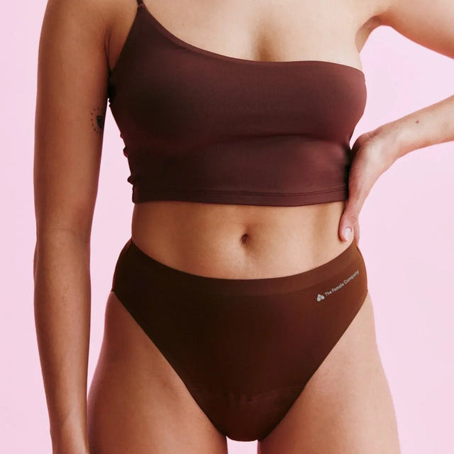 Eine Person, die ein braunes, asymmetrisches Crop-Top und dazu passende hoch taillierte Unterwäsche trägt, posiert mit einer Hand auf der Hüfte vor einem rosa Hintergrund.