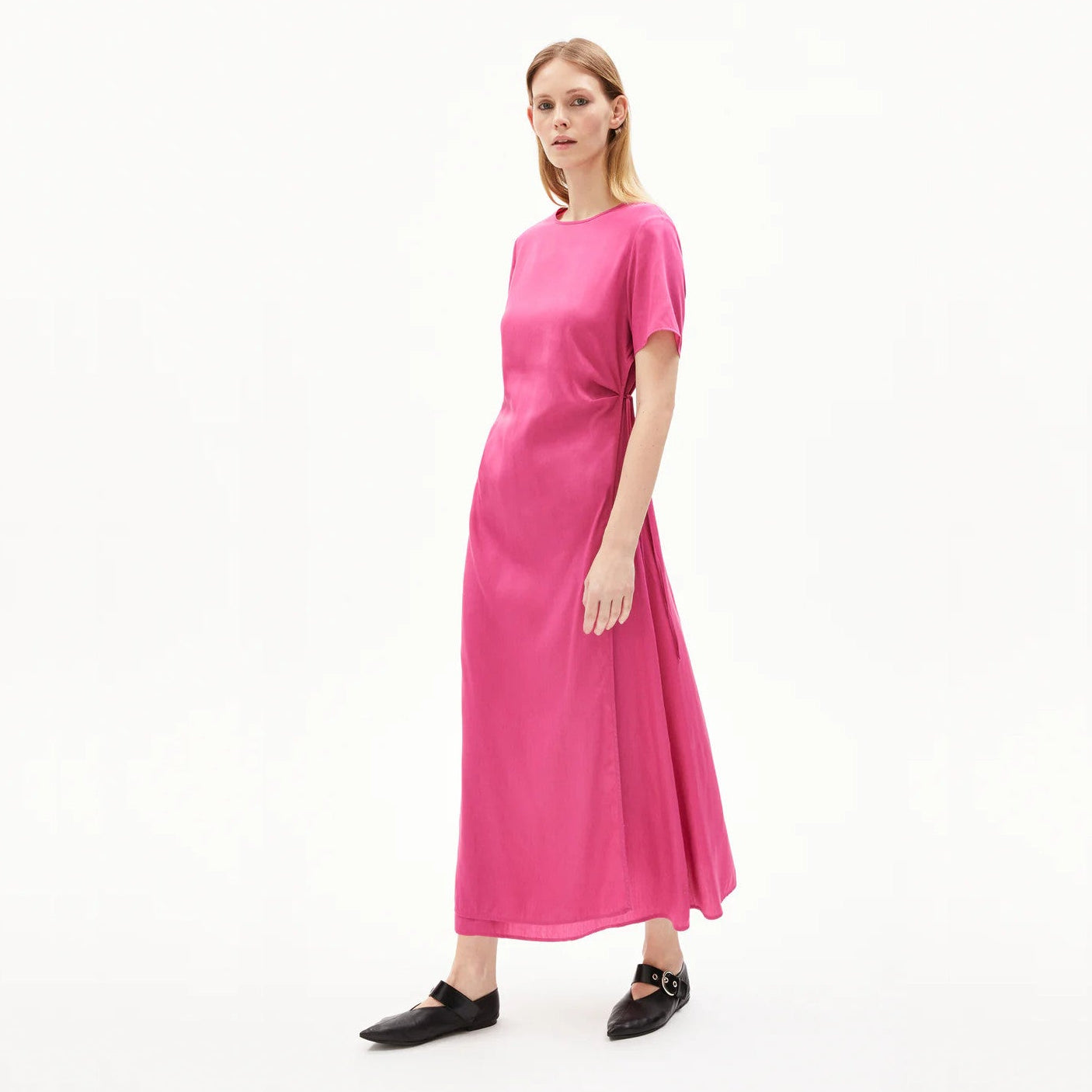 Eine Person, die vor einem schlichten weißen Hintergrund steht, trägt ein hellrosa, kurzärmeliges, knöchellanges Kleid und schwarze flache Schuhe.