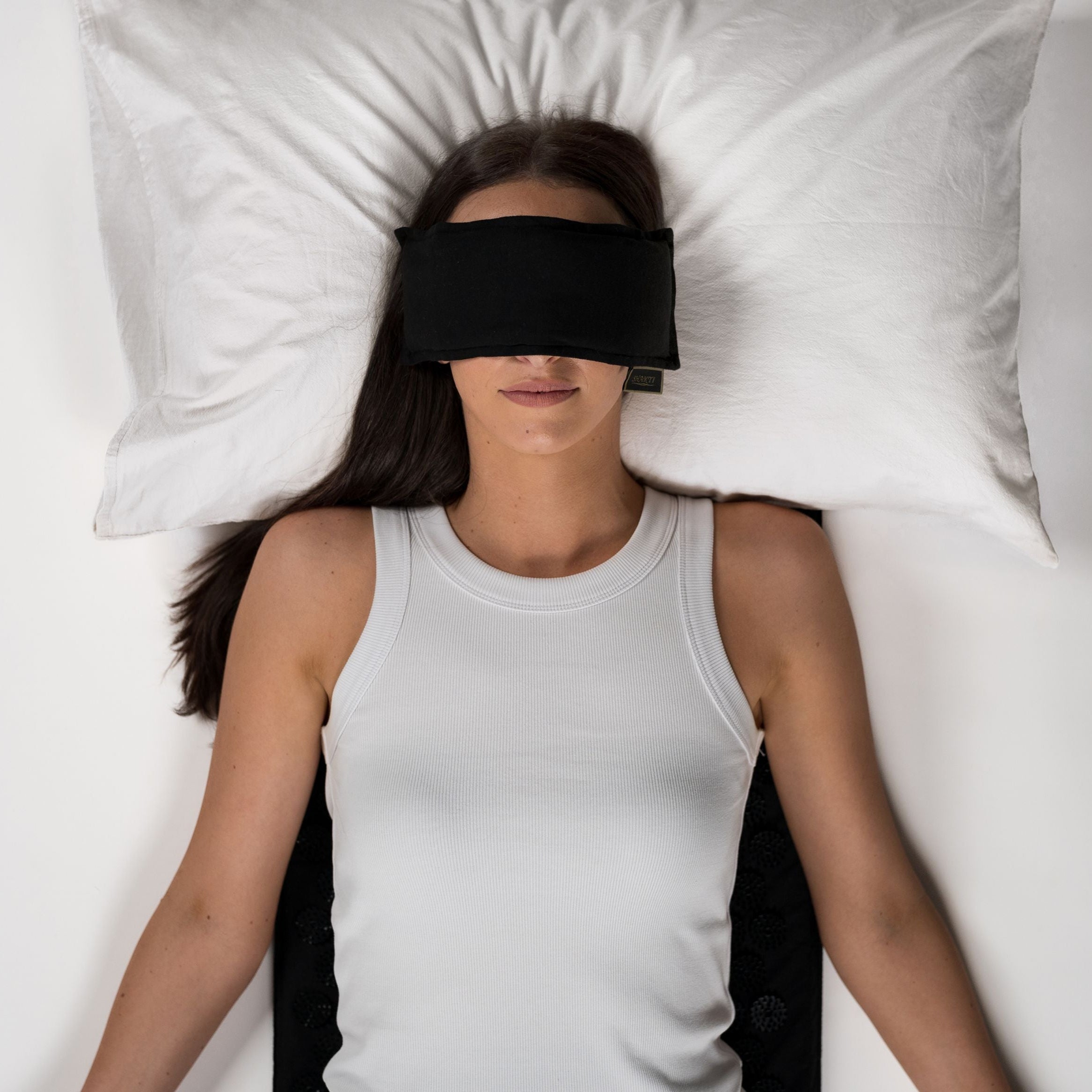 Eine Frau in einem weißen Tank-Top liegt auf dem Rücken auf einer weißen Fläche, mit einem weißen Kissen unter ihrem Kopf und einer schwarzen Augenmaske über ihren Augen. Ihre Arme ruhen an ihren Seiten.