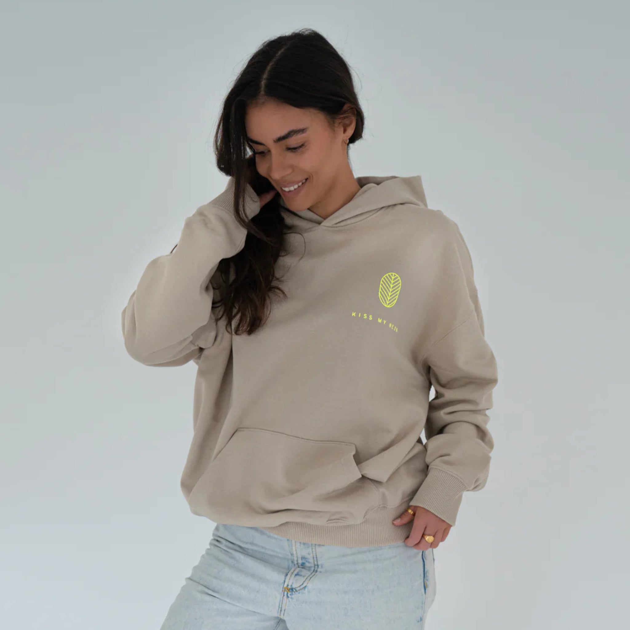 Eine Frau mit langen dunklen Haaren trägt einen beigen Kapuzenpulli mit einem kleinen Blattmuster und Text auf der Brust, gepaart mit hellblauen Jeans. Sie steht vor einem schlichten hellen Hintergrund und lächelt, während sie nach unten schaut.