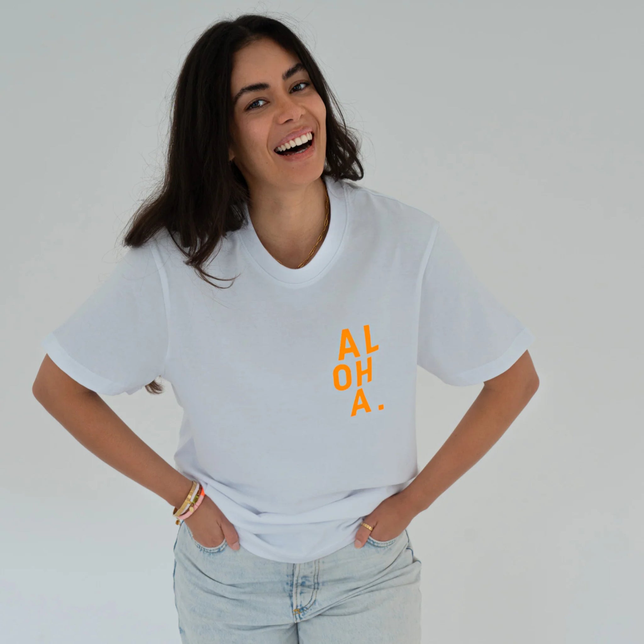 Eine lächelnde Frau mit langen dunklen Haaren trägt ein weißes T-Shirt mit der Aufschrift ALOHA. in gelb-orangenen Buchstaben auf der Brust und hellblaue Jeans vor einem schlichten hellen Hintergrund.