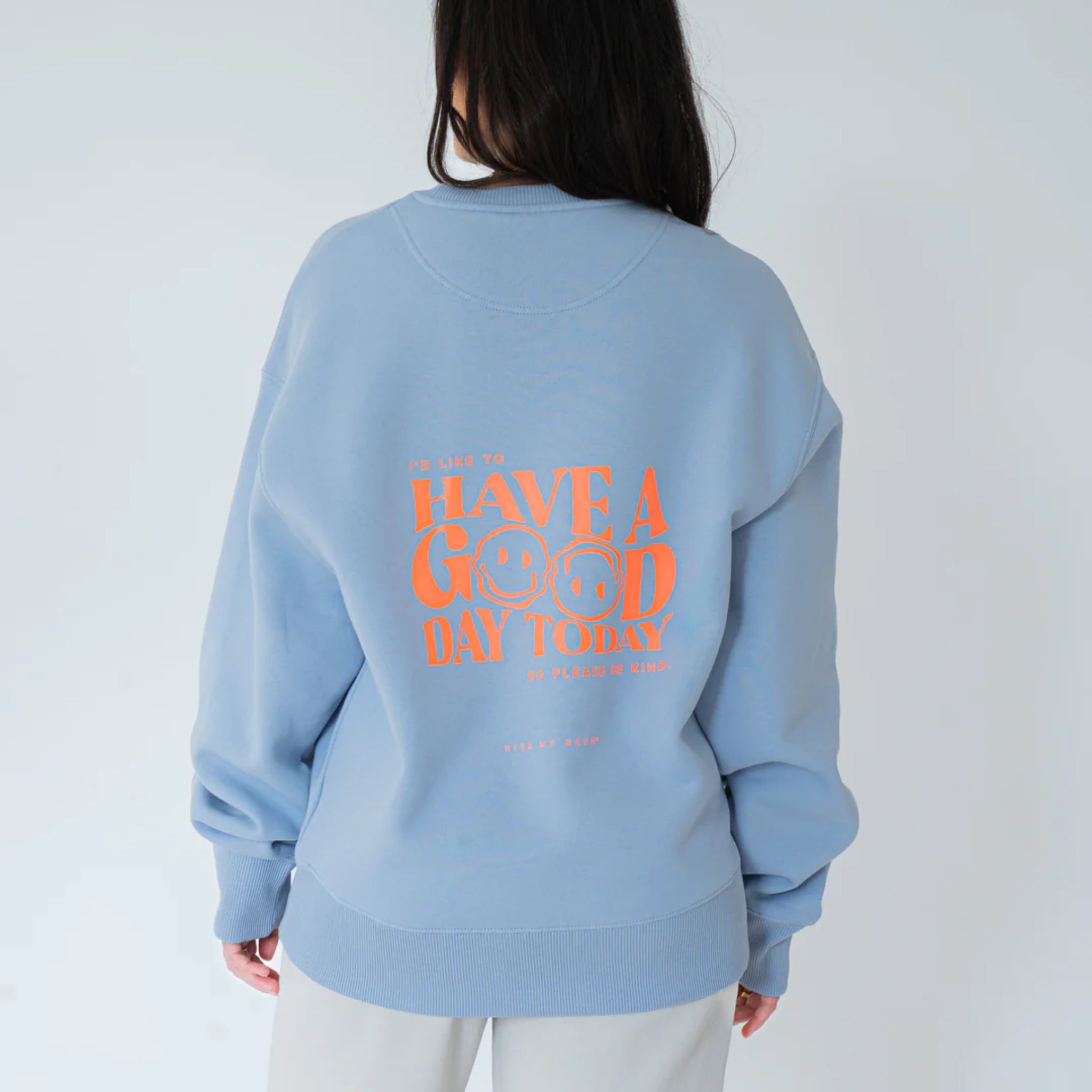 Eine Person trägt ein hellblaues Sweatshirt mit orangefarbenem Text auf der Rückseite, der lautet: "I'D LIKE TO HAVE A GOOD DAY TODAY" und zwei Smiley-Gesichter zeigt. Die Person blickt von der Kamera weg.