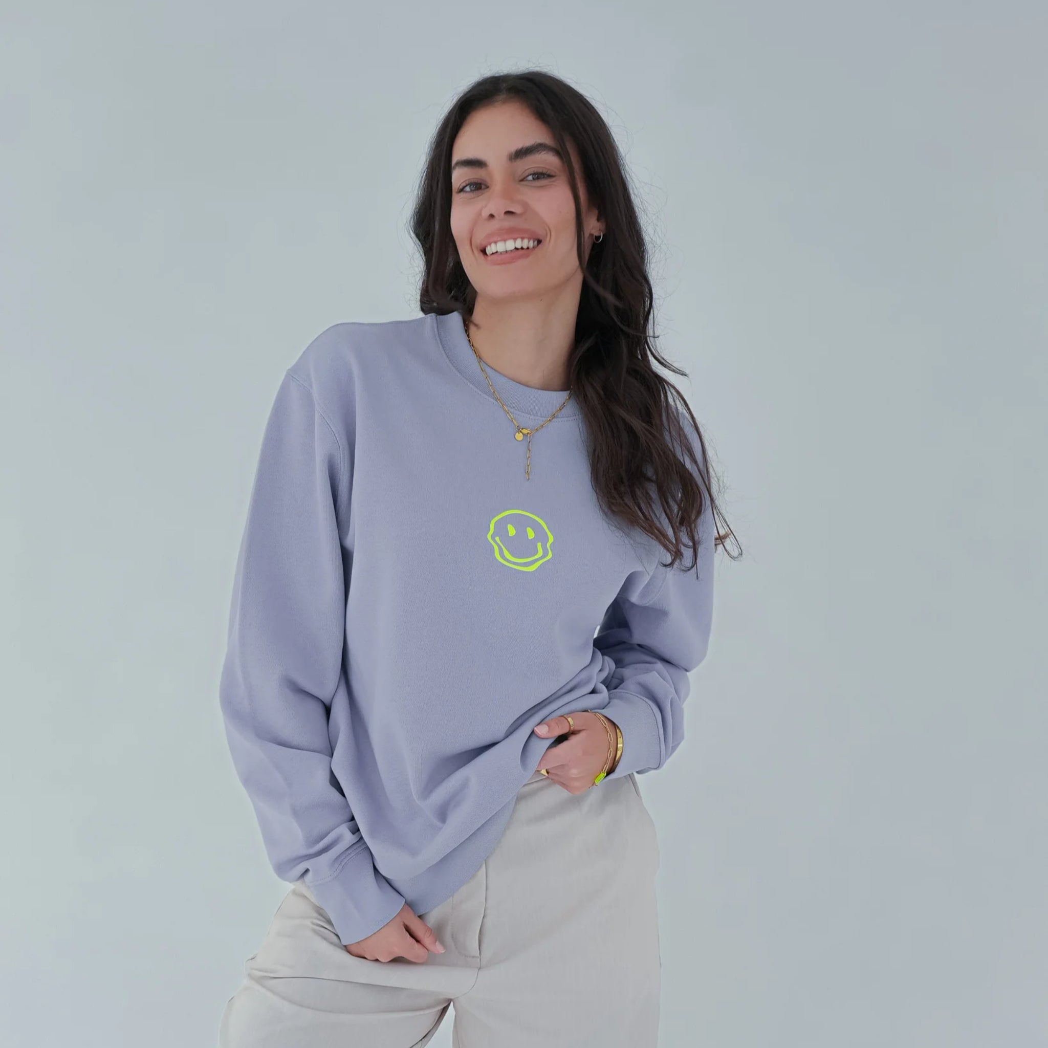 Eine Frau mit langen dunklen Haaren lächelt, während sie ein hellviolettes Sweatshirt mit einem kleinen gelben Smiley darauf, eine hellgraue Hose und Goldschmuck vor einem schlichten hellen Hintergrund trägt.