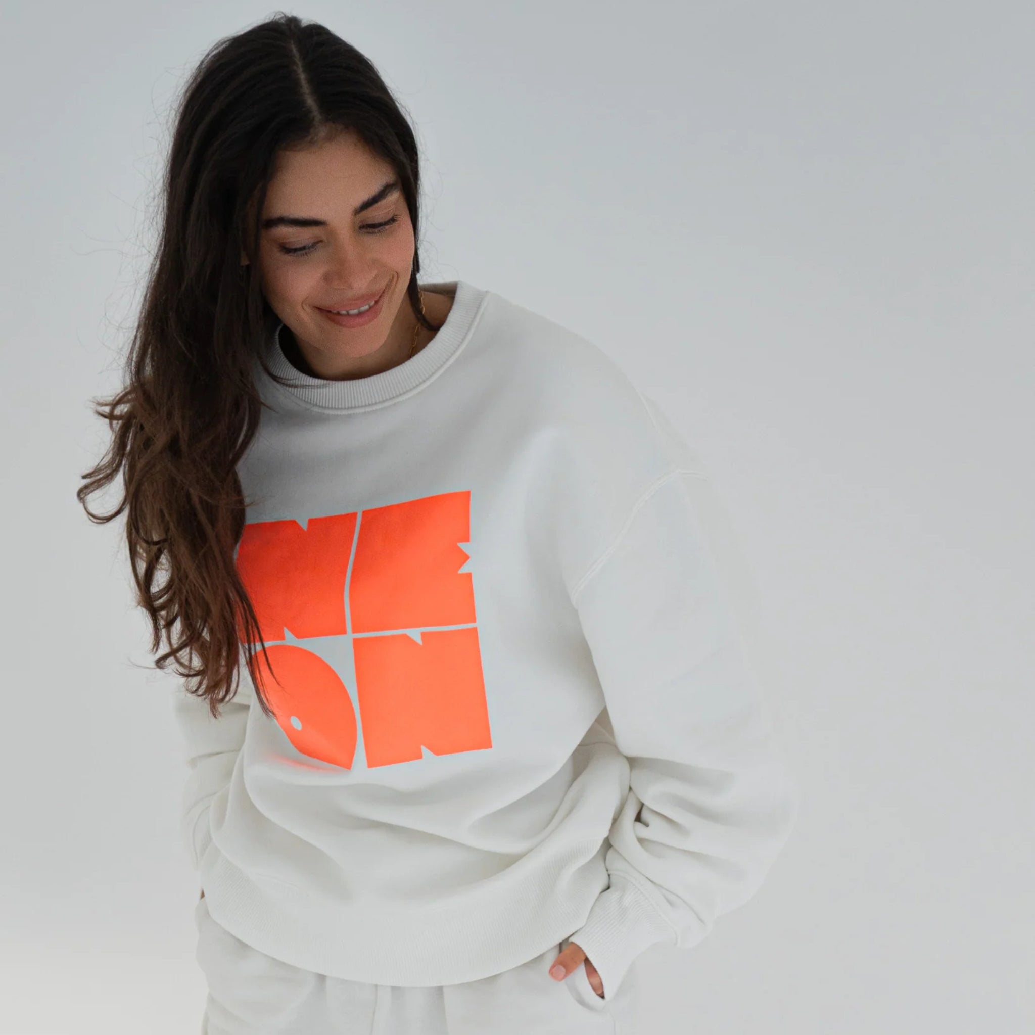 Eine Frau mit langen braunen Haaren trägt ein weißes Sweatshirt mit einem auffälligen orangefarbenen Grafikdesign. Sie blickt nach unten und lächelt, während sie vor einem schlichten hellen Hintergrund steht.