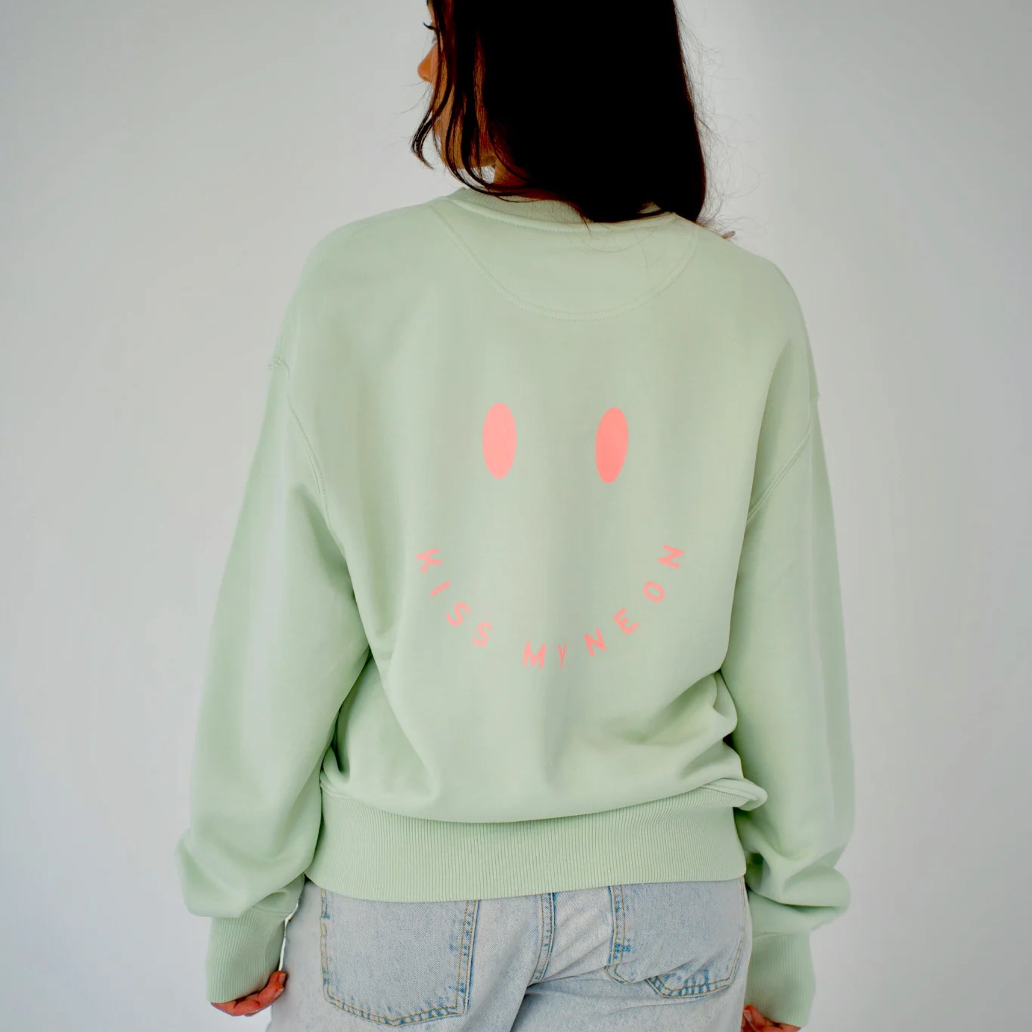 Eine Person, die ein hellgrünes Sweatshirt mit einem rosafarbenen Smiley und der Aufschrift KISS MY NEON auf dem Rücken trägt, gepaart mit einer hellblauen Jeans, steht vor einem schlichten Hintergrund.