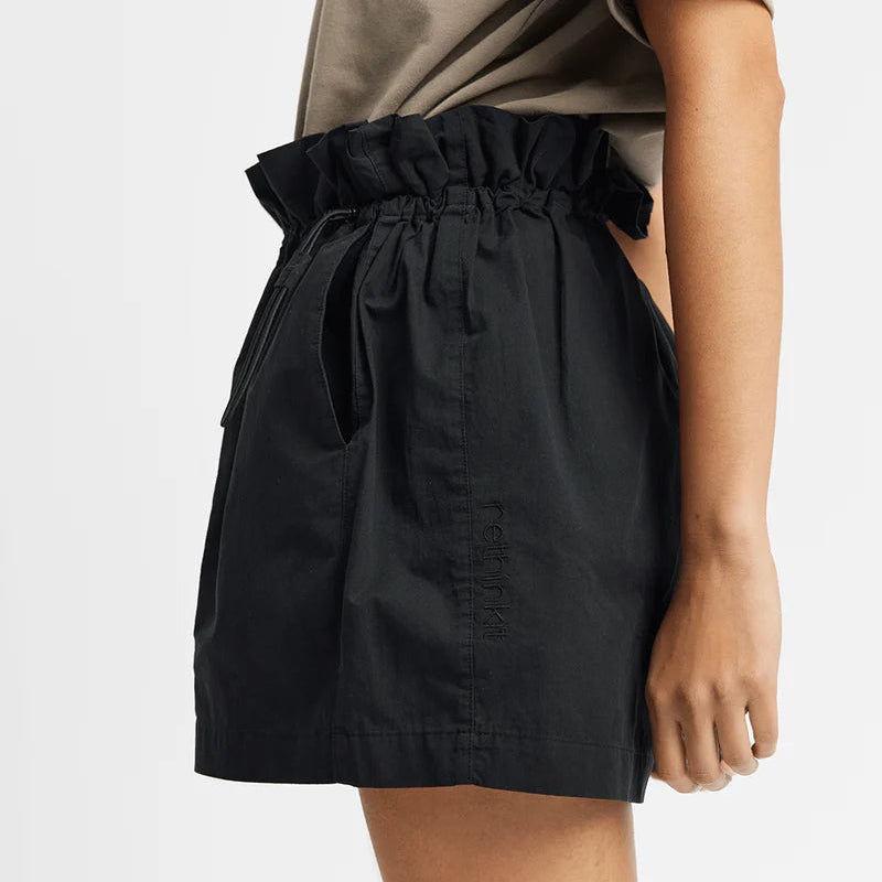 Eine Person steht im Profil und trägt hochgeschnittene schwarze Paperbag-Shorts mit einem elastischen, gerüschten Bund und ein taupefarbenes kurzärmeliges Hemd. Der Hintergrund ist einfarbig weiß.