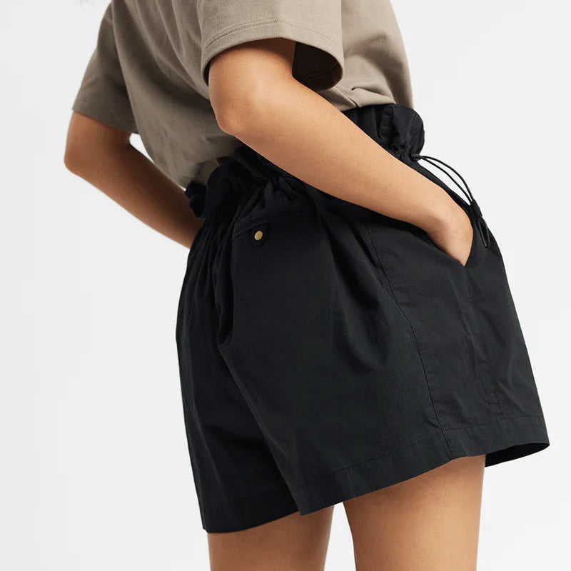 Eine Person steht mit einer Hand in der Tasche ihrer schwarzen, hochgeschlossenen Shorts und trägt ein taupefarbenes kurzärmeliges Hemd. Ihr Haar ist zu einem Zopf zusammengebunden, und der Hintergrund ist einfarbig weiß.