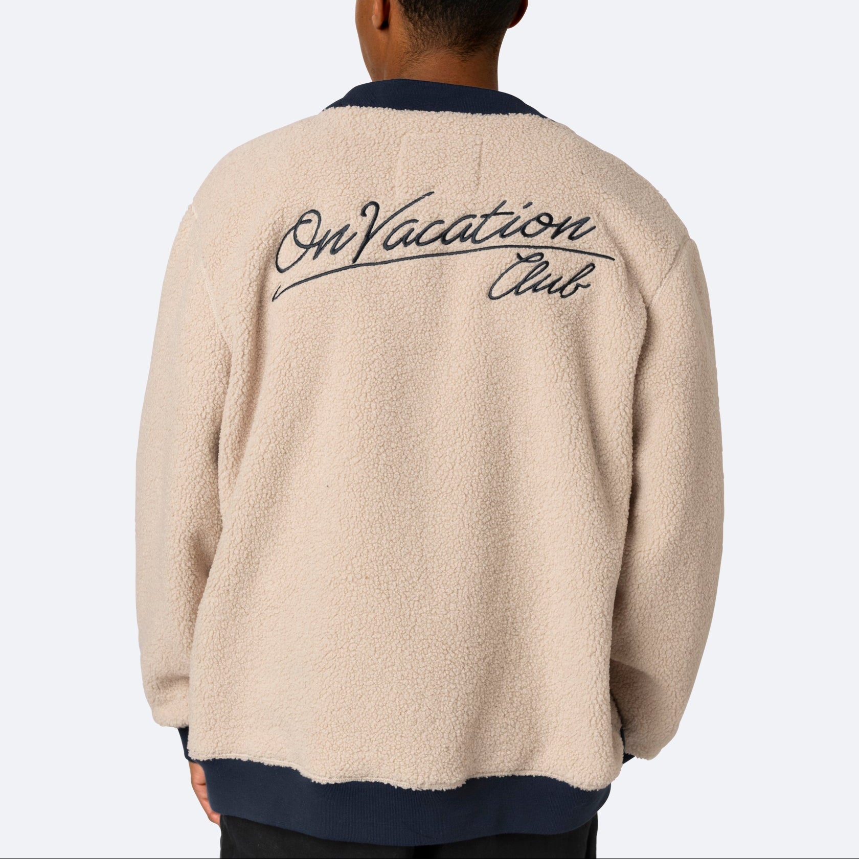 Eine Person mit Blickrichtung nach außen, die eine beigefarbene Fleecejacke trägt, auf deren Rückseite in schwarzer Schrift On Vacation Club steht. Die Jacke hat eine entspannte, gemütliche Passform und marineblaue Kragen Details.