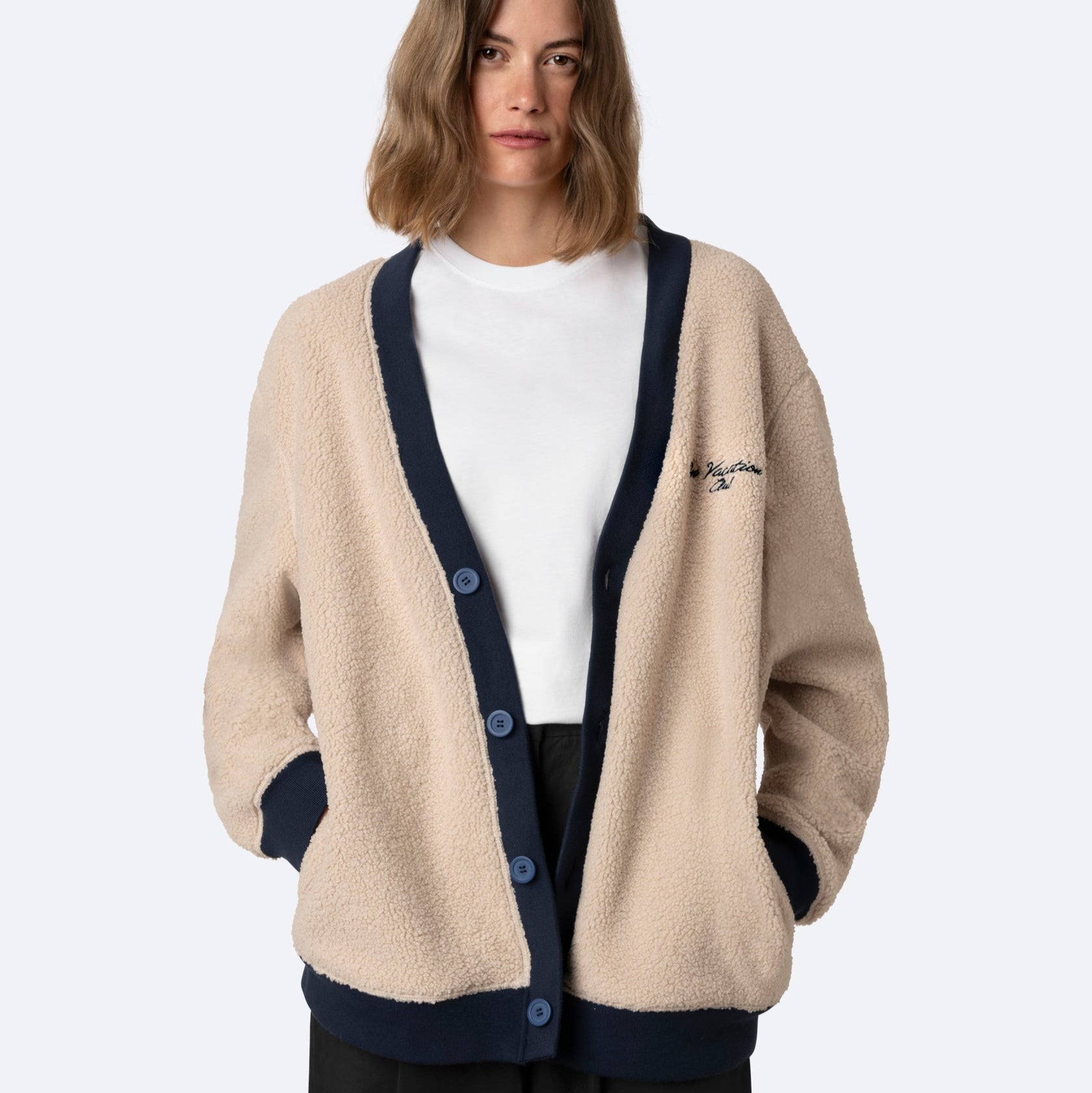 Frau mit schulterlangem gewelltem Haar trägt eine beigefarbene Fleece-Strickjacke mit marineblauem Saum und Knöpfen über einem schlichten weißen T-Shirt und einer schwarzen Hose vor einem hellgrauen Hintergrund.