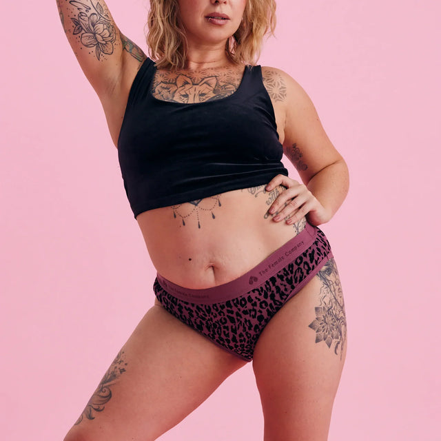 Eine Frau mit schulterlangen blonden Haaren und Tattoos posiert selbstbewusst vor einem rosa Hintergrund. Sie trägt ein schwarzes Crop-Top und lila Unterwäsche mit Leopardenmuster.