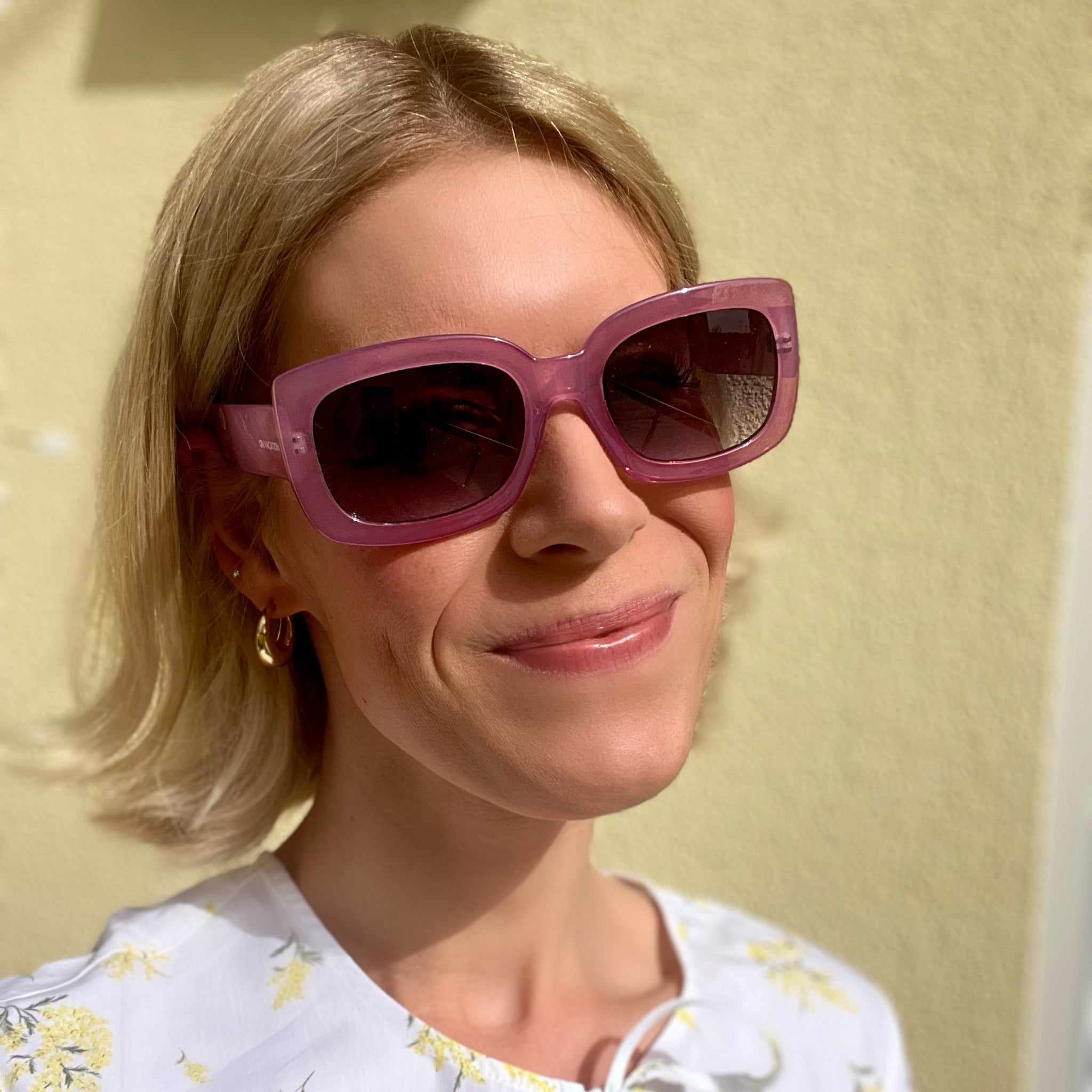Eine Frau mit blonden Haaren trägt eine große, rosafarbene Sonnenbrille, ein weiß-gelbes Oberteil und lächelt.