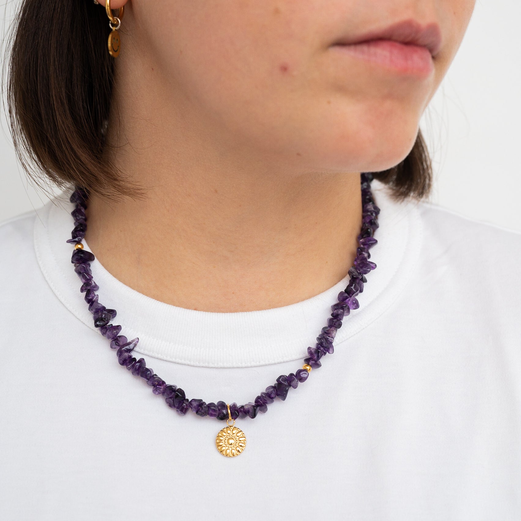 Eine Person, die eine lilafarbene Perlenkette mit einem goldenen Sonnenanhänger und einem passenden goldenen Ohrring trägt, von den Schultern aufwärts dargestellt, bekleidet mit einem schlichten weißen Hemd.