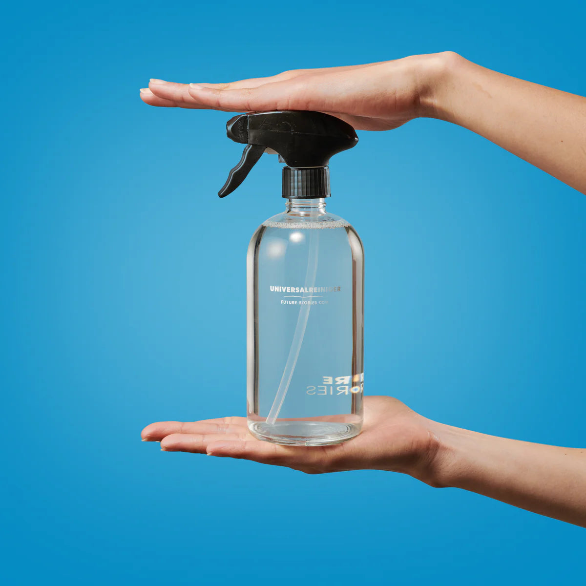 Eine durchsichtige Sprühflasche mit einer schwarzen Düse wird horizontal zwischen zwei Händen vor einem einfarbig blauen Hintergrund gehalten.