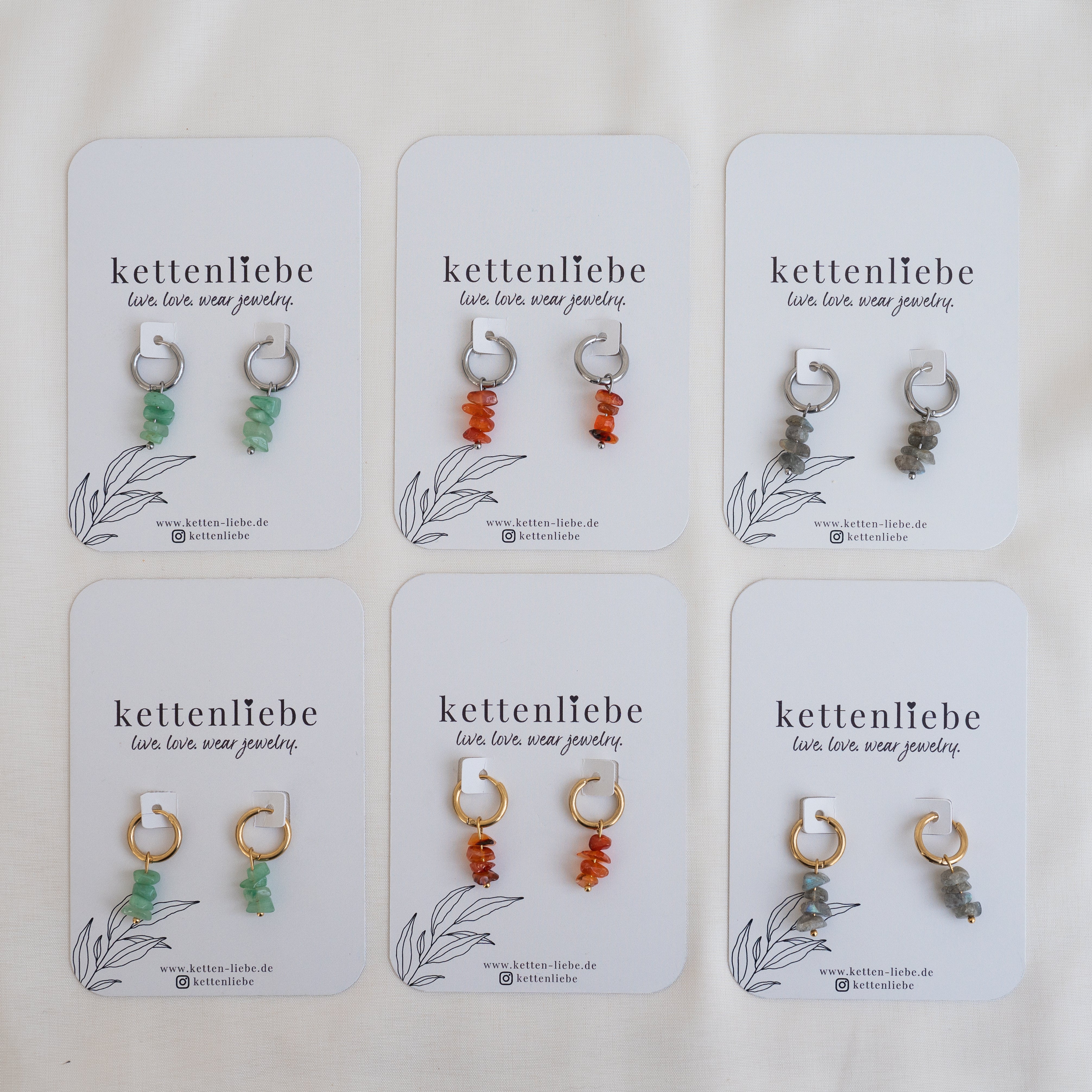 Sechs Paar Perlenohrringe in den Farben Grün, Orange und Grau sind auf Karten der Marke "kettenliebe" in zwei Dreierreihen angeordnet. Auf jeder Karte steht der Slogan "live, love, wear jewelry".