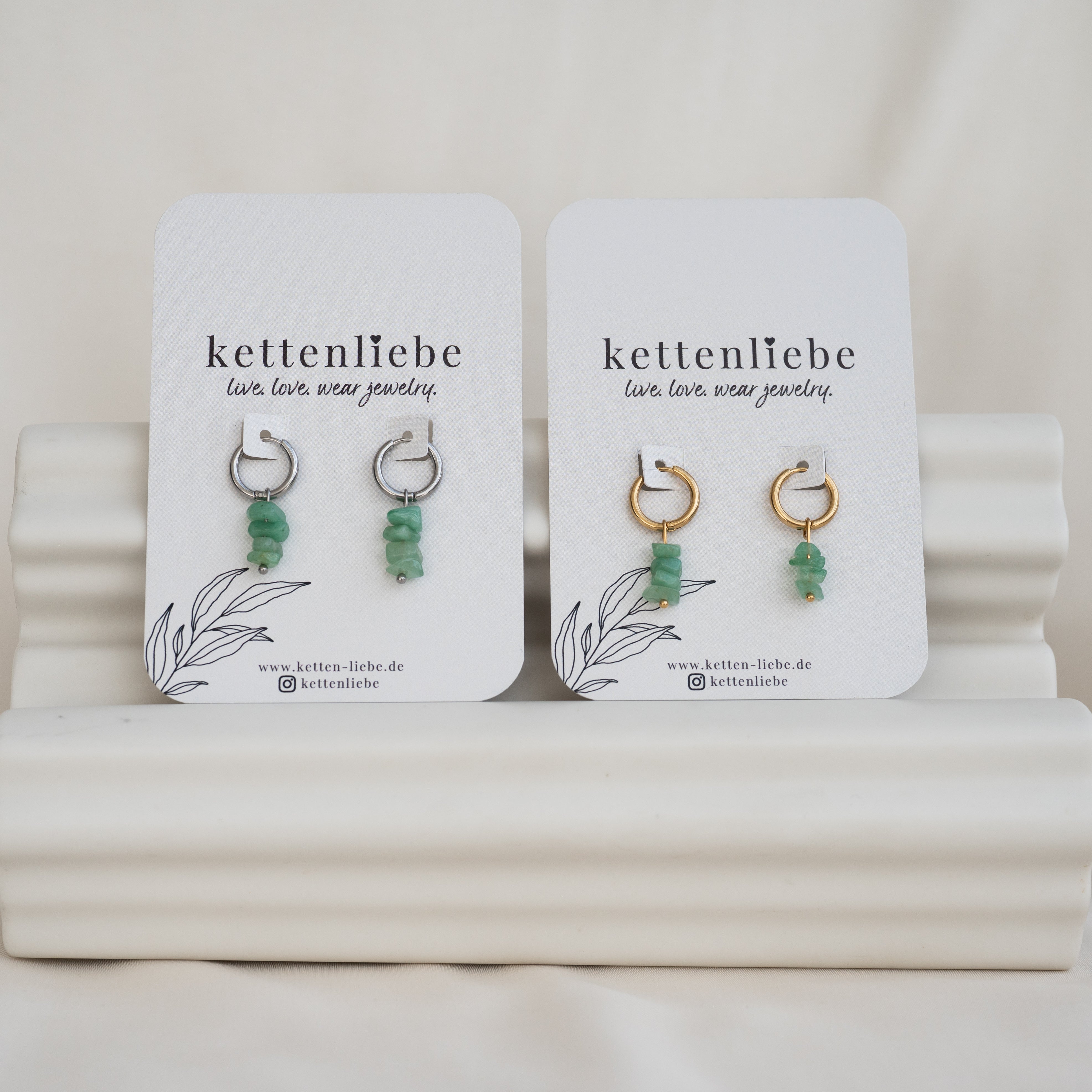 Zwei Paar Ohrringe mit grünen Steinperlen, eines mit silbernen und eines mit goldenen Reifen, auf weißen Karten mit der Aufschrift kettenliebe auf einem weißen, gewellten Ständer.