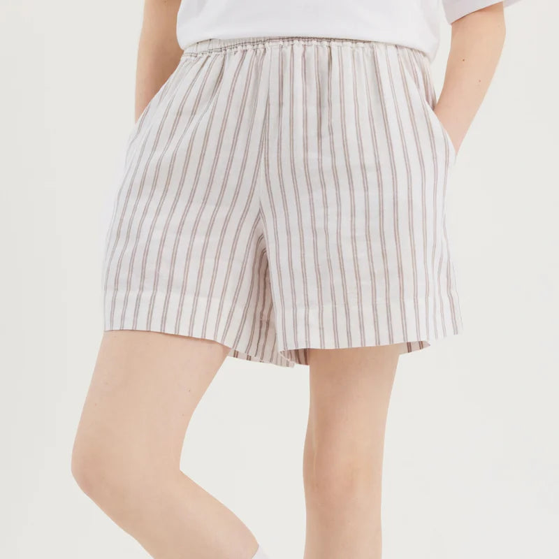 Eine Person, die ein weißes T-Shirt, beige und weiß vertikal gestreifte Shorts, weiße Socken und weiße Turnschuhe mit beigen Details trägt, steht mit den Händen in den Taschen vor einem schlichten Hintergrund.
