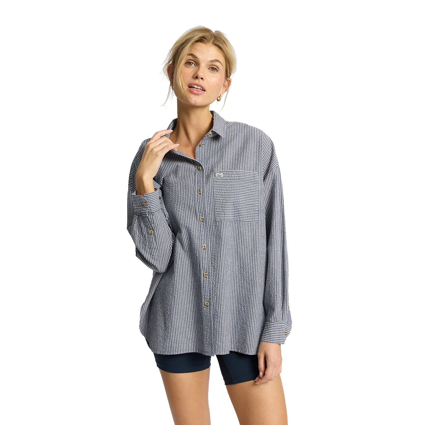 Eine Frau, die ein locker sitzendes, blau-weiß gestreiftes Button-up-Hemd mit einer Vordertasche und marineblaue Shorts trägt, steht vor einem schlichten weißen Hintergrund.