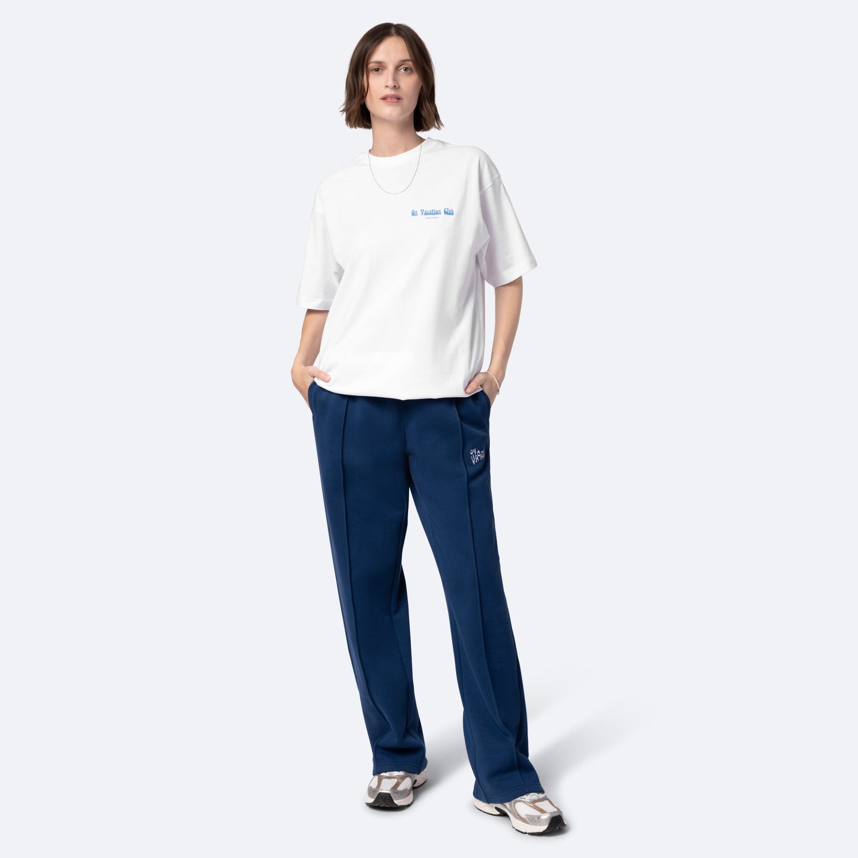 Eine Person steht mit dem Gesicht nach vorne und trägt ein lockeres weißes T-Shirt, eine blaue Hose mit weitem Bein und Turnschuhe. Sie hat kurzes braunes Haar und einen neutralen Gesichtsausdruck, die Hände in den Taschen, vor einem schlichten hellen Hintergrund.
