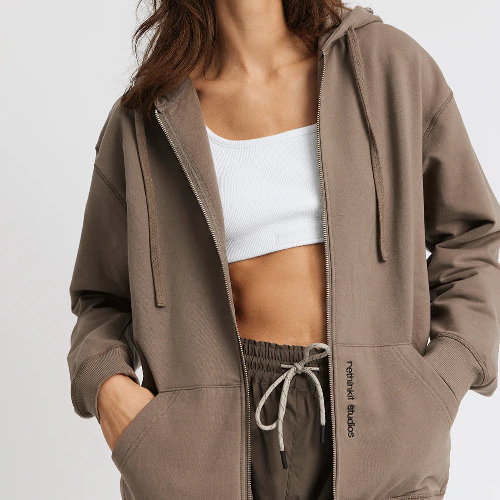 Frau in einer braunen Kapuzenjacke, weißem Top und brauner Hose mit Kordelzug, Hände in den Taschen, von vorn fotografiert.