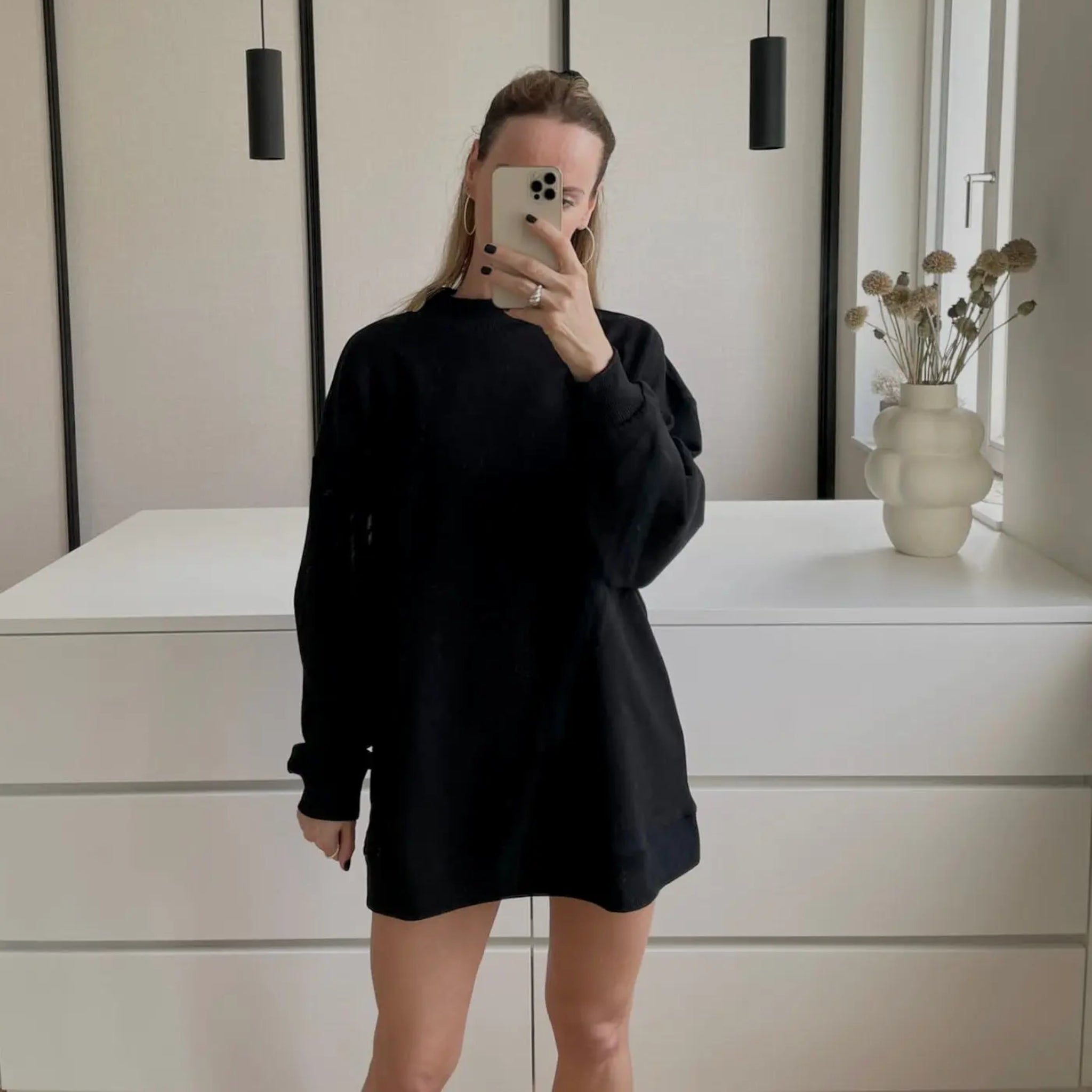 Eine Person in einem lockeren schwarzen Sweatshirt-Kleid steht in einem Haus und hält sich ein Telefon vor das Gesicht. Hinter ihr steht eine weiße Kommode mit einer modernen Vase mit Trockenblumen und zwei Hängelampen.