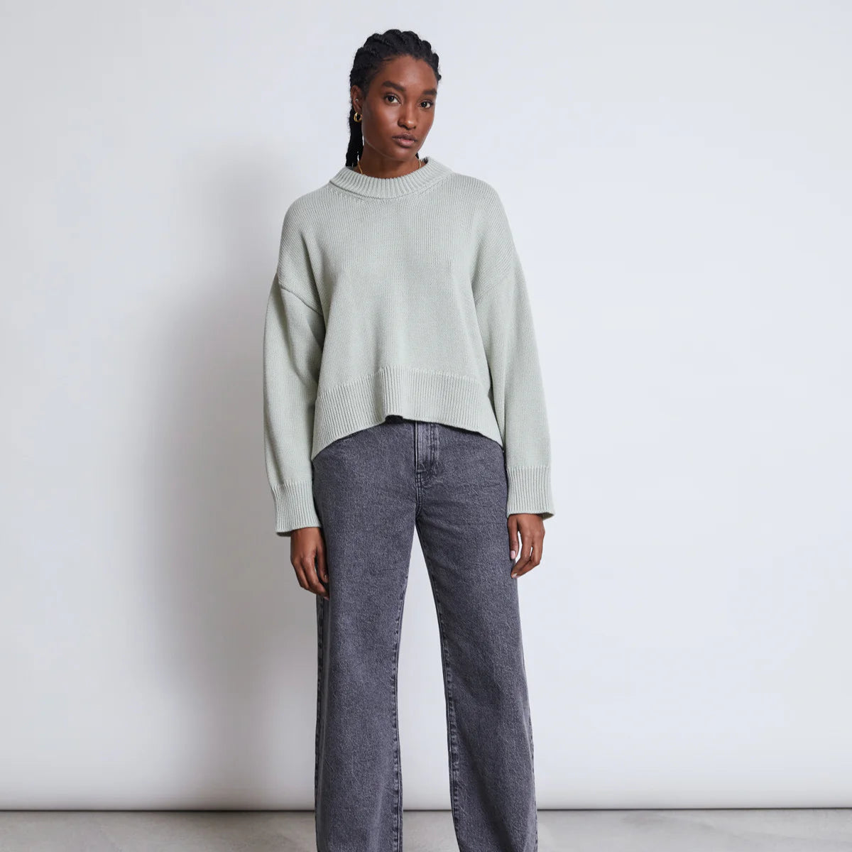 Eine Frau steht vor einer schlichten hellen Wand und trägt einen lockeren hellgrauen Pullover, dunkelgraue Jeans mit weitem Bein und schwarze Schuhe. Sie hat geflochtenes Haar und einen neutralen Gesichtsausdruck.