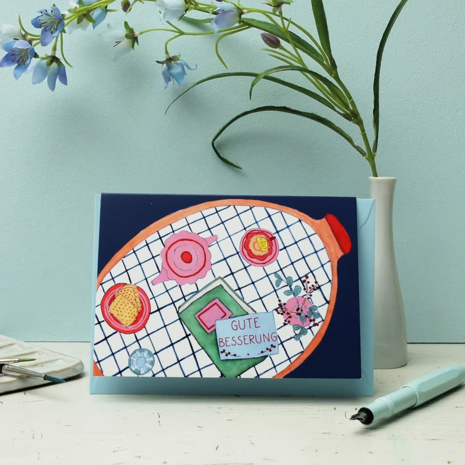 Eine Karte "Gute Besserung" mit bunten Abbildungen von Blumen, Tee, Honig, Zitrone und einer Wärmflasche steht auf einem hellblauen Tisch neben einer Vase mit kleinen lila Blumen und einem Stift.