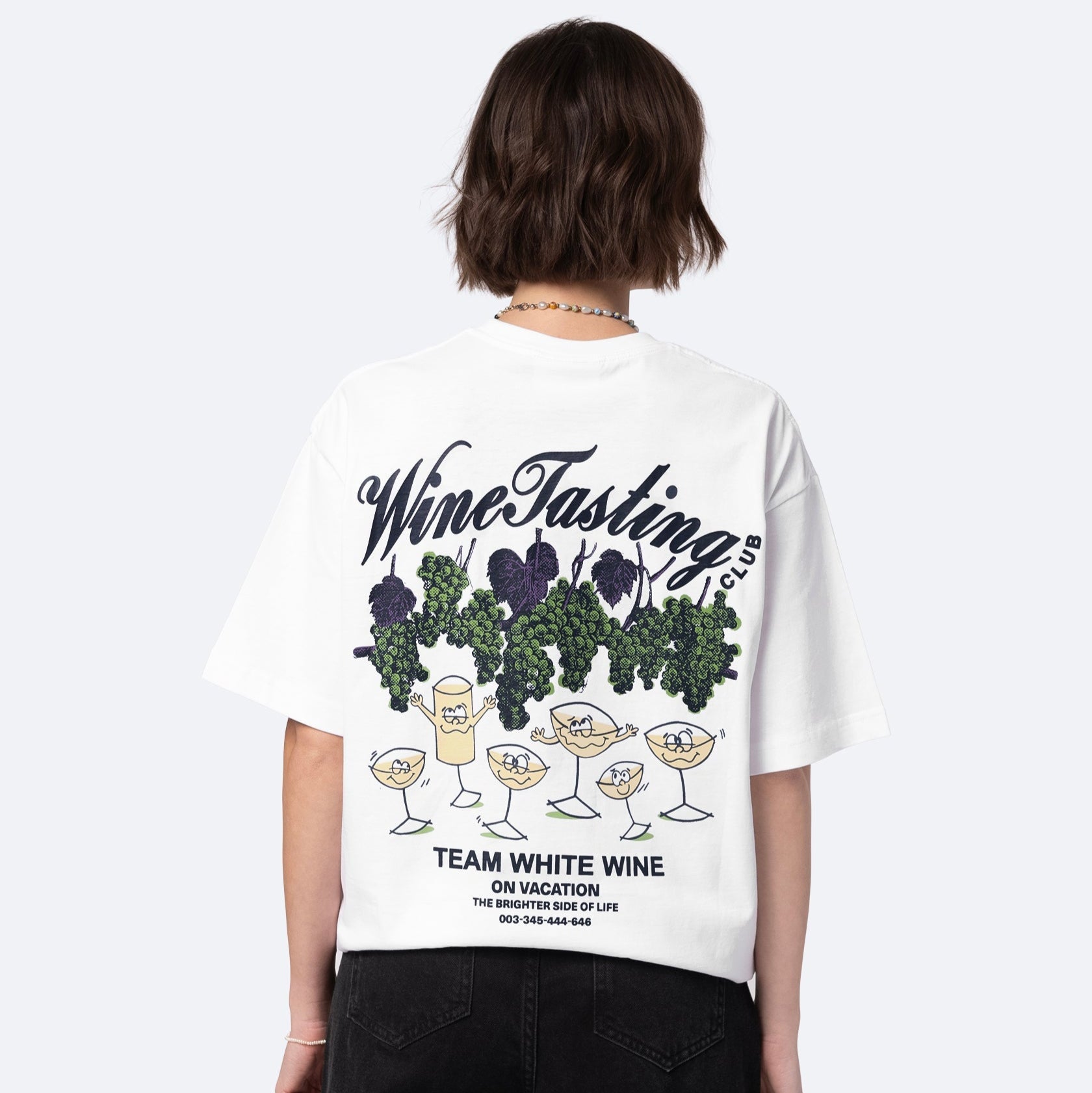 Person, die ein weißes T-Shirt mit einem illustrierten Design von Weingläsern, Trauben und Blättern trägt. Auf dem T-Shirt steht "Wine Tasting Club, Team White Wine", und es enthält zusätzlichen spielerischen Text und eine Telefonnummer.