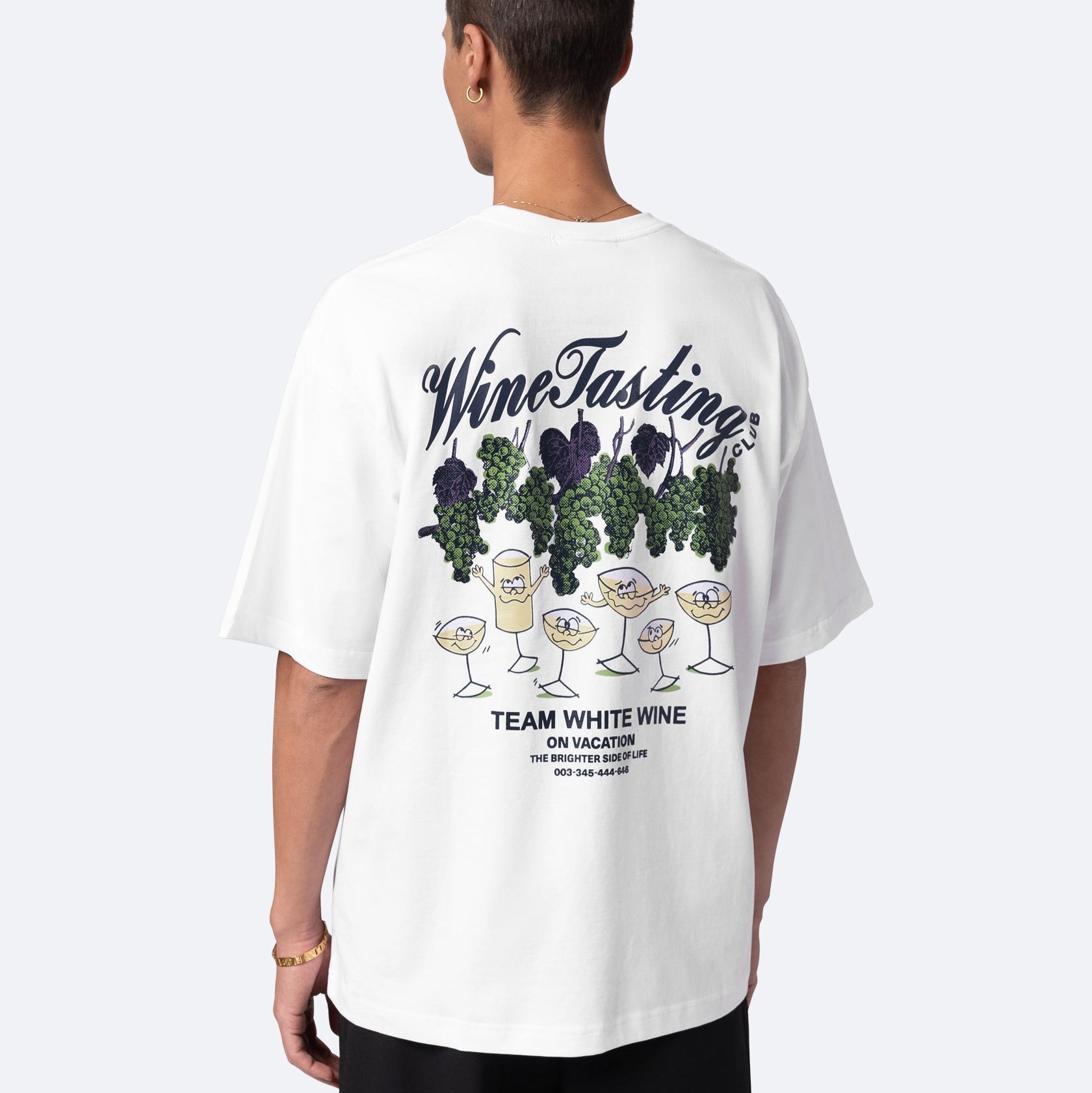 Eine Person, die von hinten gesehen wird und ein weißes T-Shirt mit einem illustrierten Muster aus Weingläsern, Trauben und Blättern trägt. Der Text auf dem T-Shirt lautet "Wine Tasting", "Team White Wine on Vacation" und ein weiterer kleiner Text darunter.
