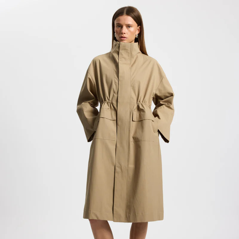 Eine Frau, die einen langen, hellbraunen Trenchcoat mit hohem Kragen und großen Vordertaschen trägt, steht vor einem schlichten weißen Hintergrund und hat die Hände in den Manteltaschen.