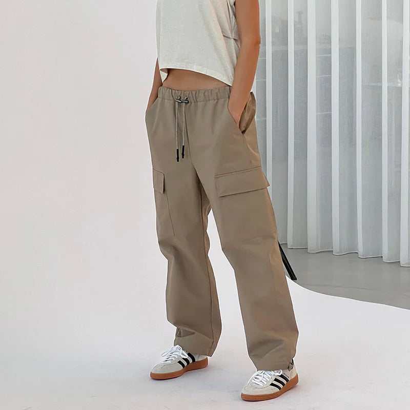 Eine Frau steht in einem minimalistischen Studio und trägt ein ärmelloses weißes Tanktop, eine weite beigefarbene Cargohose und schwarz-weiße Turnschuhe. Sie hat die Hände in den Taschen und blickt zur Seite. Im Hintergrund sind weiße vertikale Jalousien zu sehen.