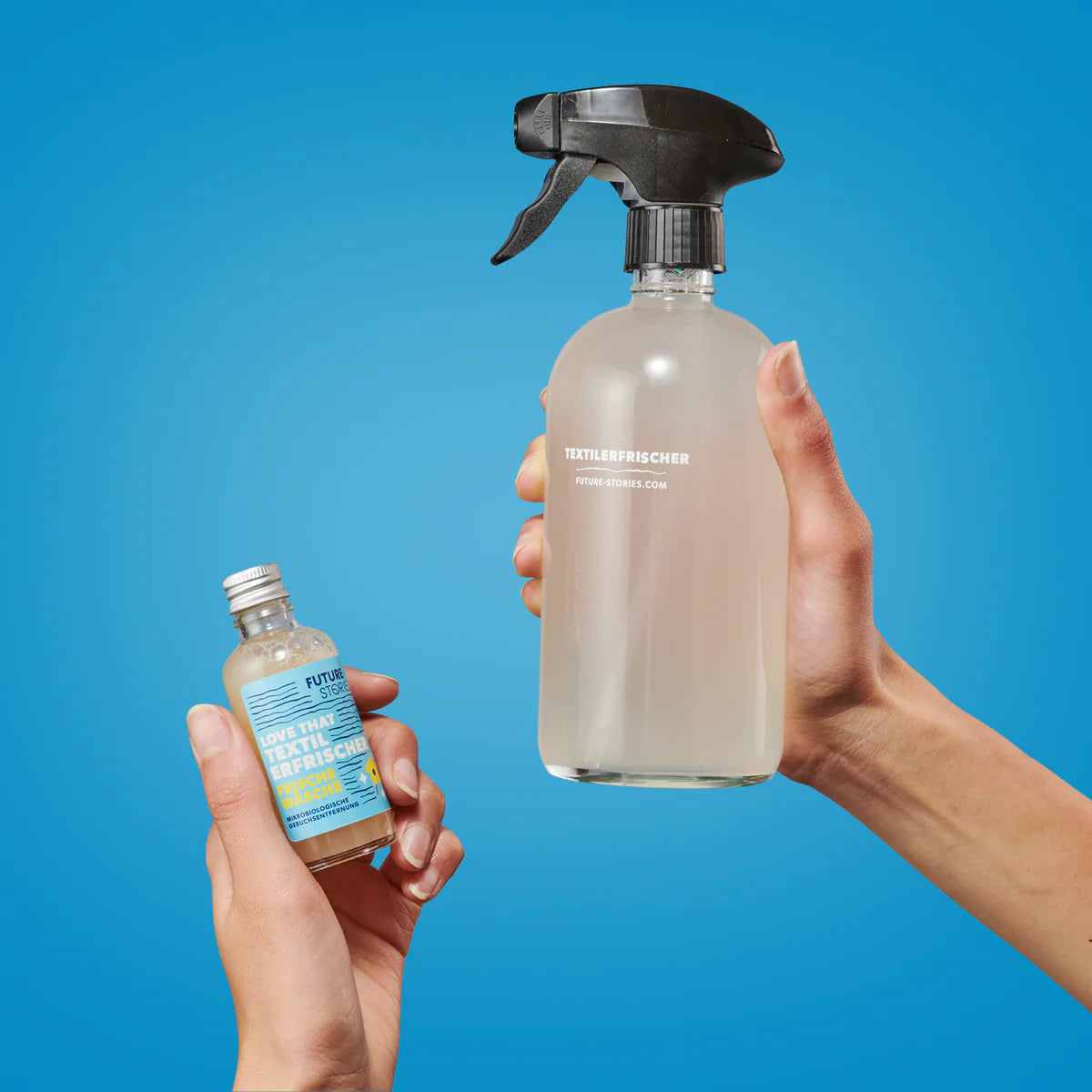 Zwei Hände halten Reinigungsmittel vor einem blauen Hintergrund: eine hält eine kleine Flasche mit blauem Etikett, die andere eine größere klare Sprühflasche mit der Aufschrift TEXTILERFRISCHER.