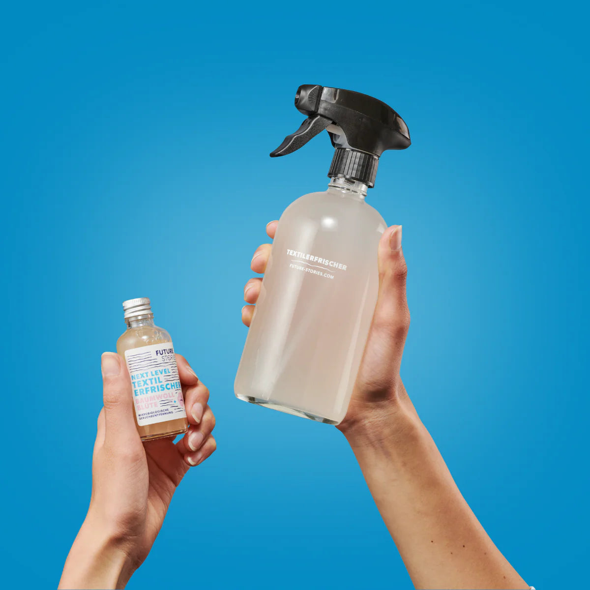 Zwei Hände halten Flaschen vor einem blauen Hintergrund. Eine Hand hält eine kleine Nachfüllflasche, die andere eine größere Sprühflasche mit der Aufschrift "Textilerfrischer".