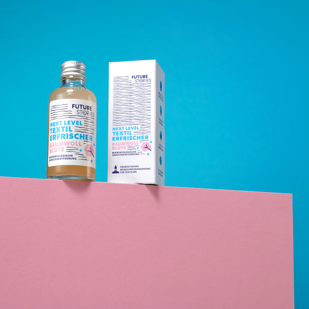 Eine Flasche und eine Schachtel Future Stories Textile Refresher stehen auf einer rosa Fläche vor einem blauen Hintergrund. Beide Artikel sind mit minimalistischen weißen und blauen Etiketten mit Text und kleinen grafischen Elementen versehen.