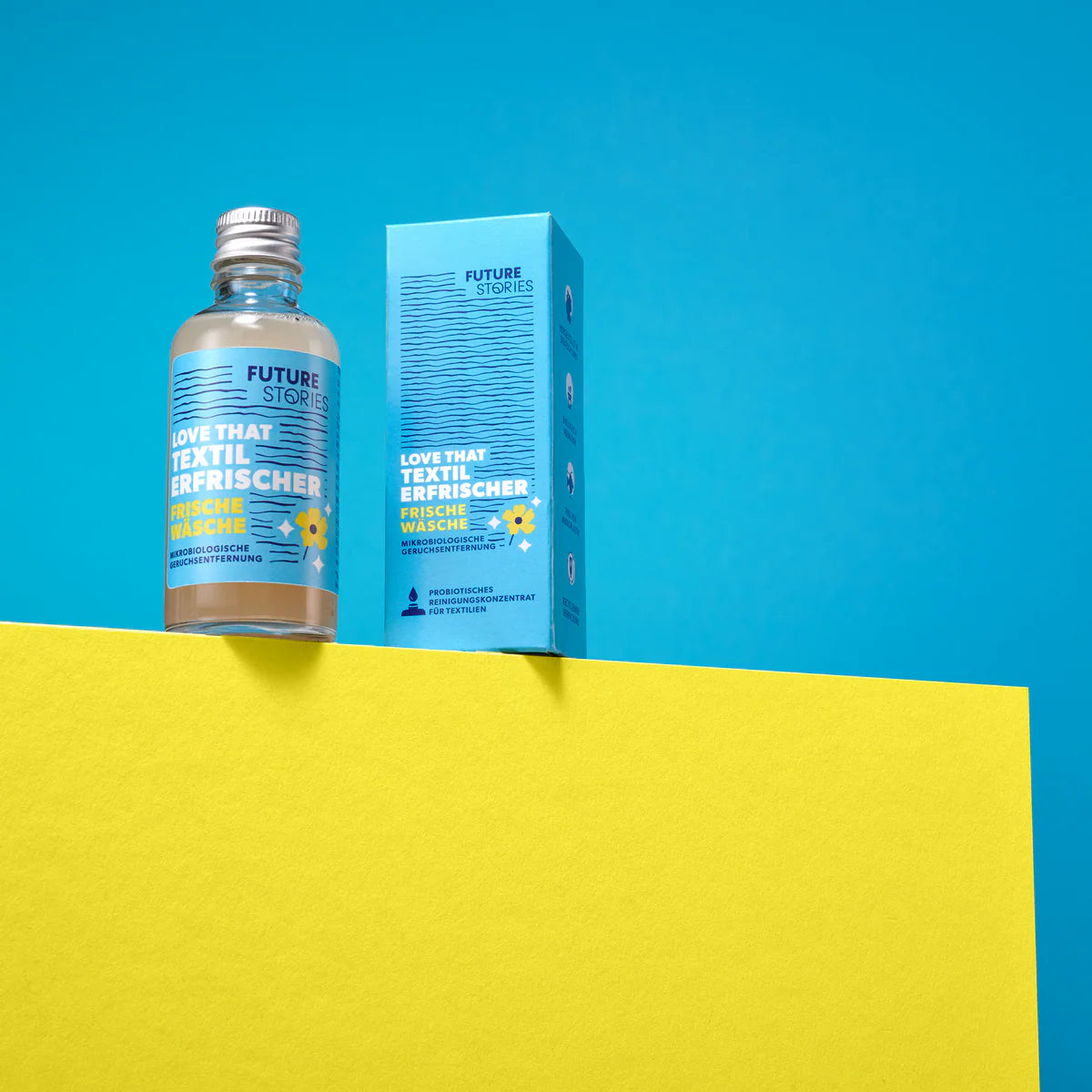 Ein kleiner Flakon und die dazugehörige Schachtel mit der Aufschrift Future Stories Love That Textil Erfrischer sind auf einer gelben Fläche vor einem blauen Hintergrund abgebildet. Die Verpackung ist blau mit gelber und weißer Schrift.