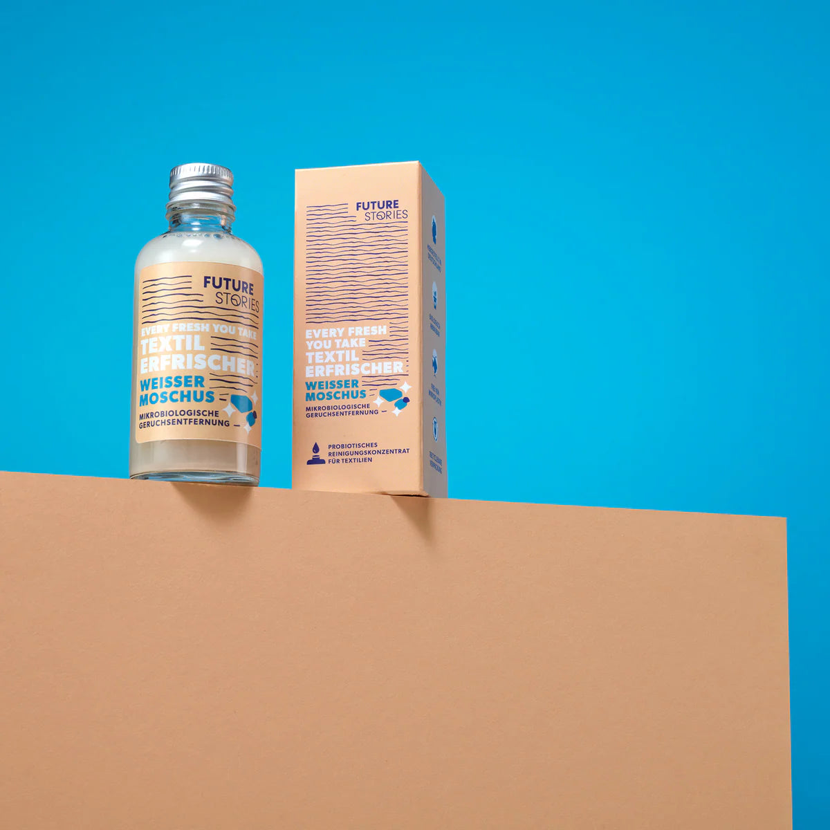 Eine Flasche und eine Schachtel von Future Stories Textilerfrischer sind auf einer beigen Fläche vor einem hellblauen Hintergrund abgebildet. Die Verpackung weist metallische und blaue Akzente mit deutschem und englischem Text auf.