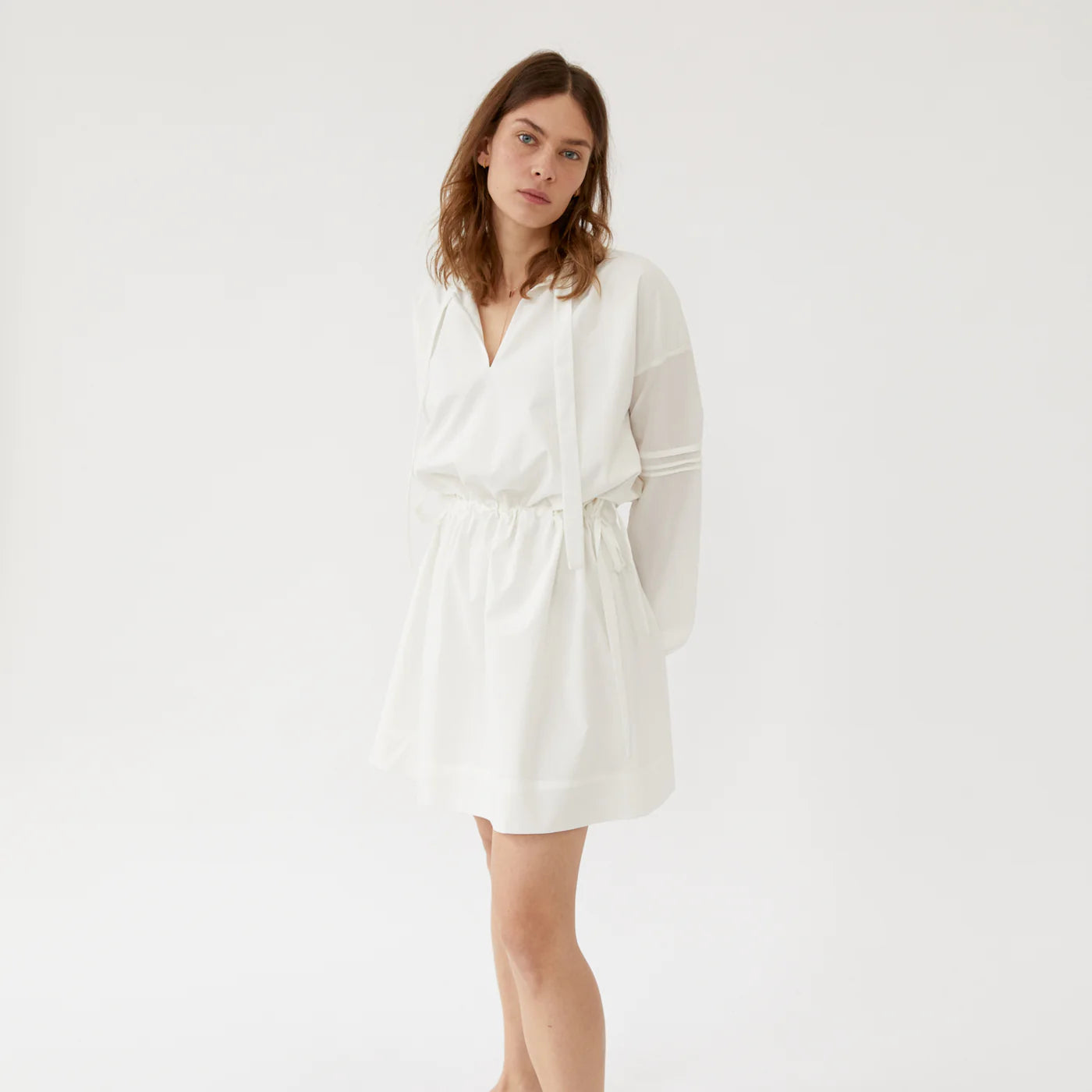 Frau in einem weißen, locker sitzenden Kleid mit langen Ärmeln vor einem neutralen Hintergrund.